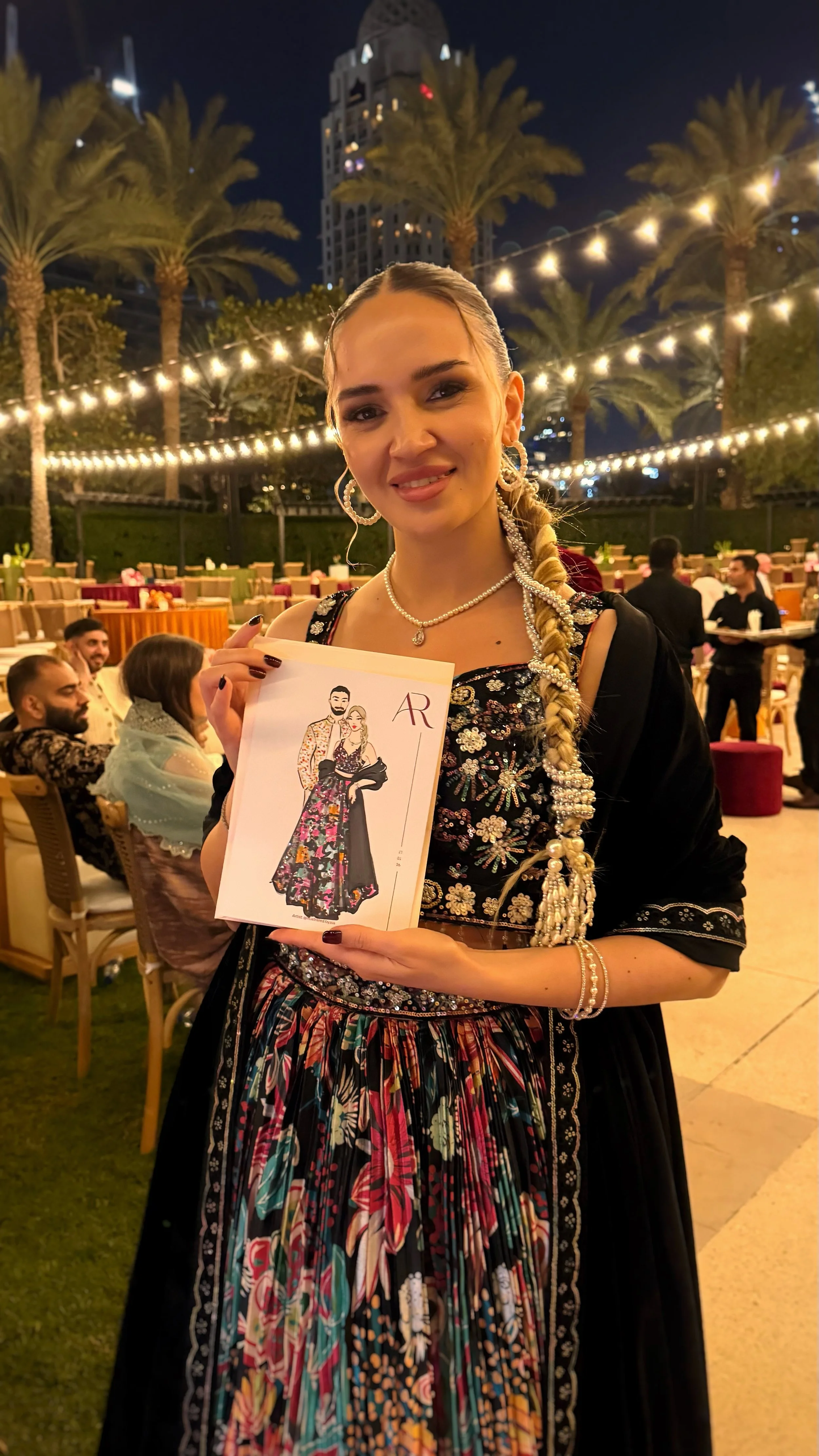 A woman in traditional attire holding a booklet with a fashion illustration, at an outdoor event during the night with string lights and palm trees in the background.