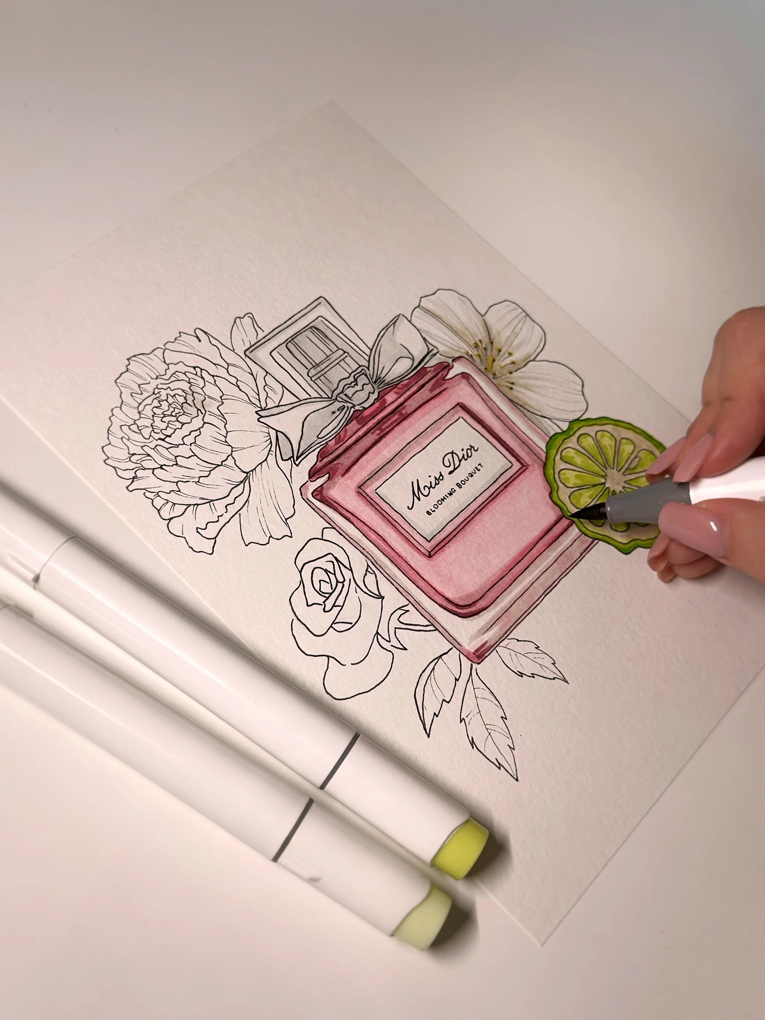 A hand coloring the floral perfume bottle illustration on paper, with visible markers and a floral-themed perfume label reading 'Moi Dior Blooming Bouquet'.
