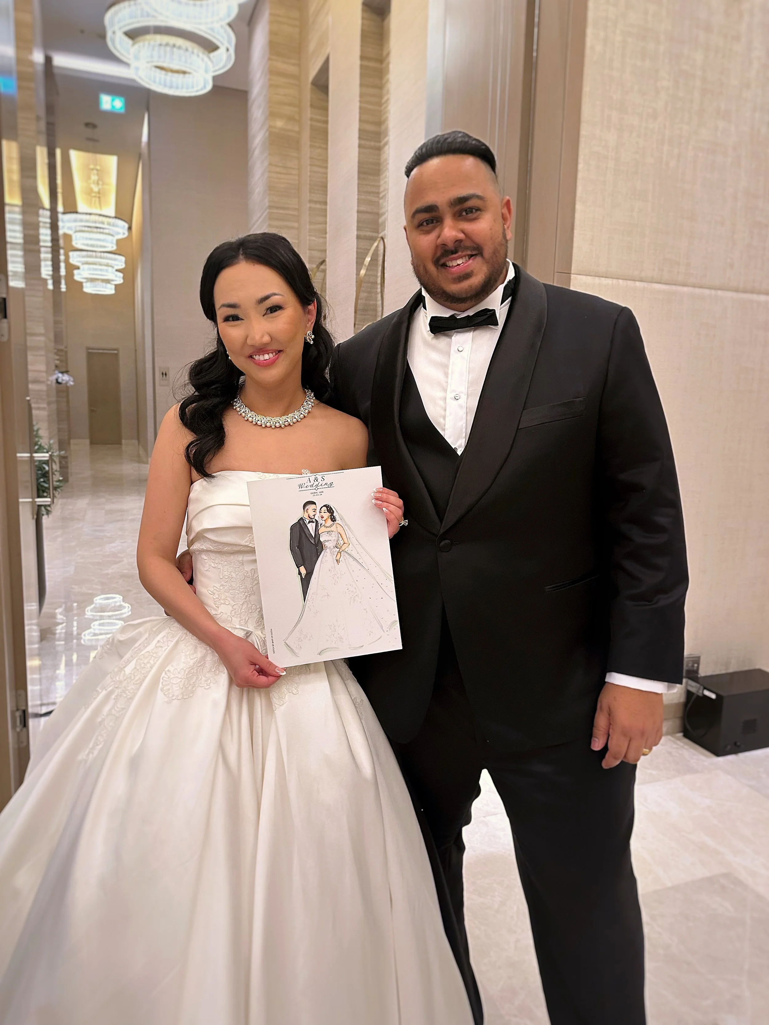 A happy bride and groom in wedding attire standing together in a fancy restaurant or hotel lobby, with the bride holding a wedding illustration. 