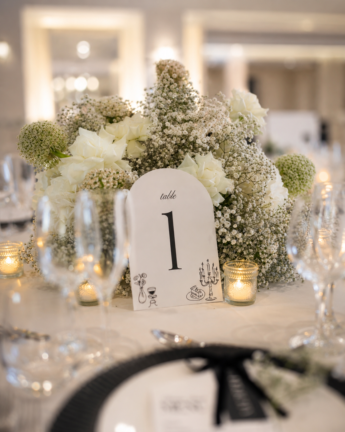 A wedding reception table decorated with a floral centerpiece of white flowers and candles, with a table number card marked '1' surrounded by glassware.