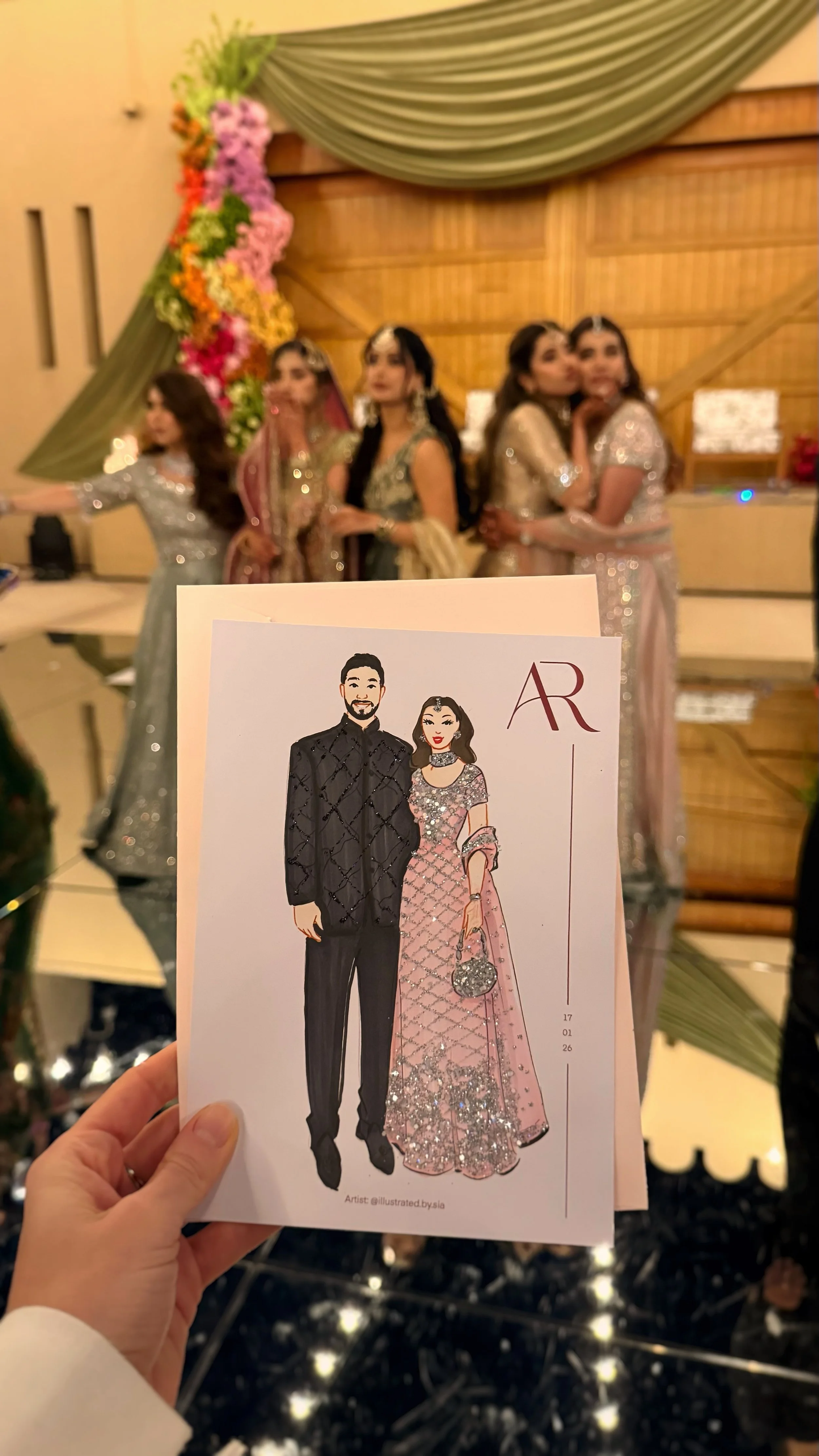 A hand holding a wedding invitation with a cartoon illustration of a couple in traditional attire. In the background, four women in elegant dresses pose for a photo at a decorated event.