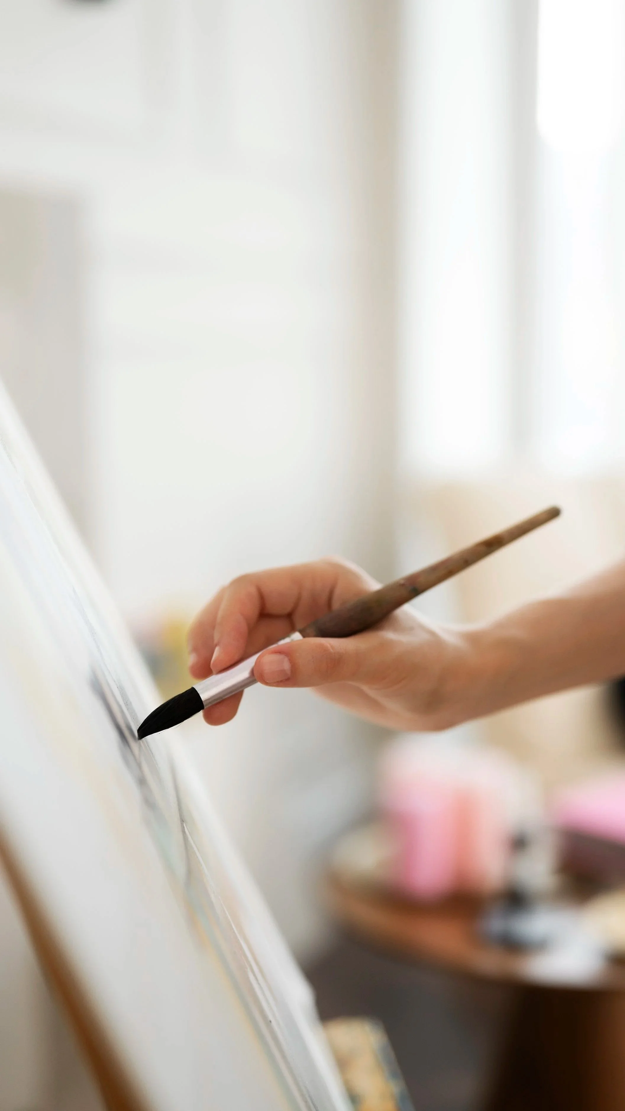 Close-up of a person's hand holding a black paintbrush, painting on a white canvas or wall in a bright room.