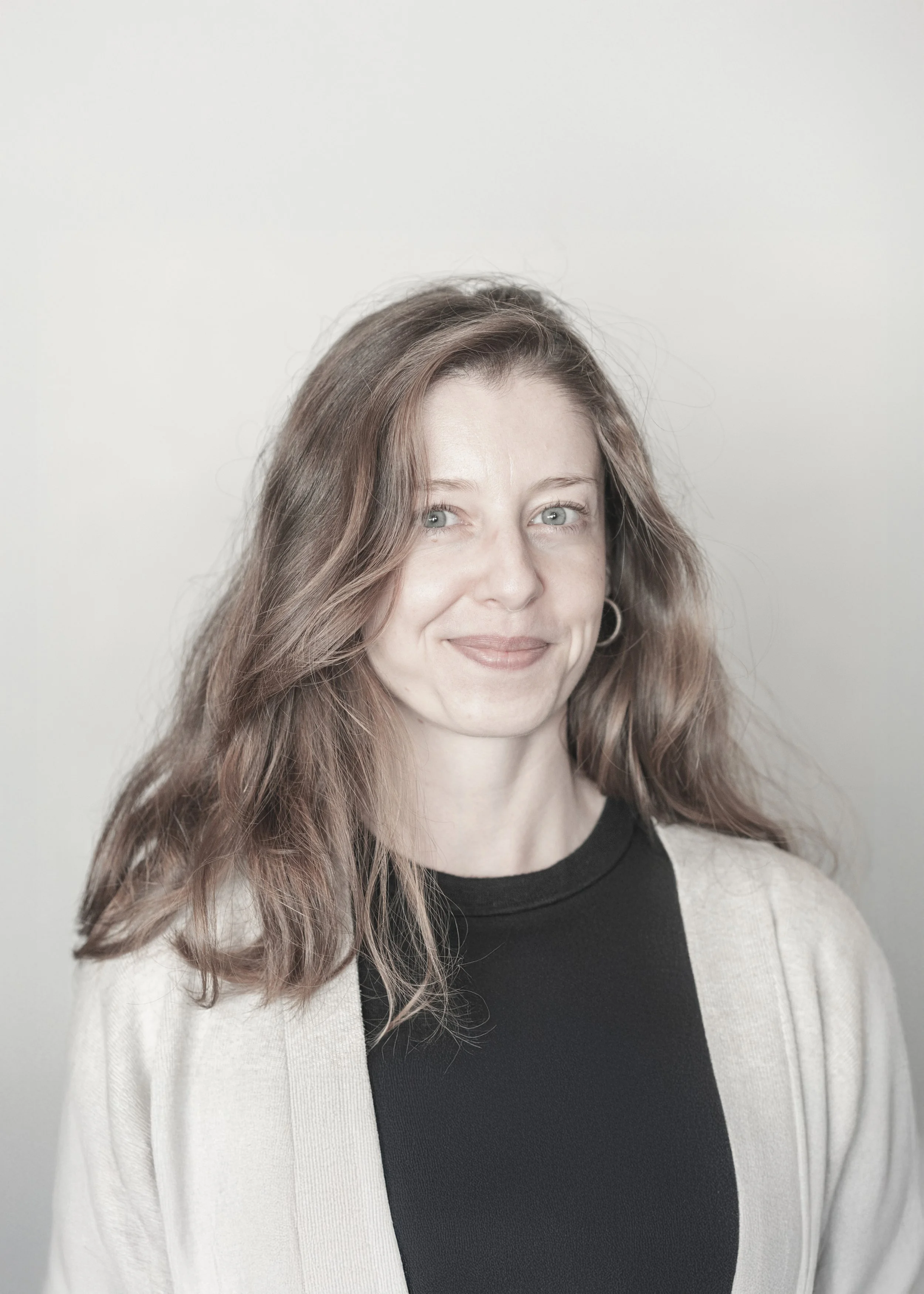 A woman with long wavy brown hair and blue eyes, wearing a black top and a beige cardigan, smiling softly against a plain light background.