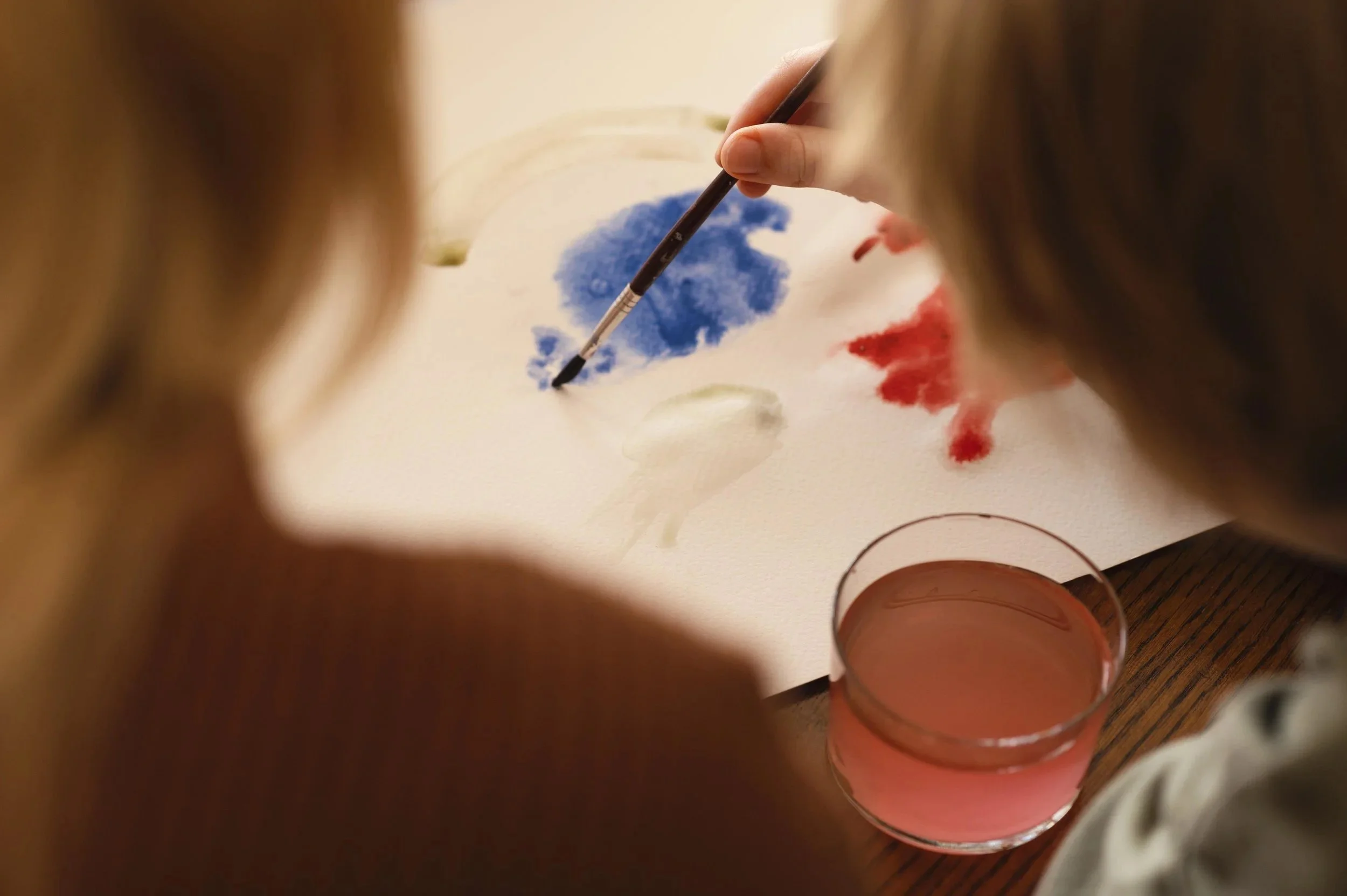 One child painting with watercolors on paper, one using a brush with blue paint, and a glass of pinkish water.