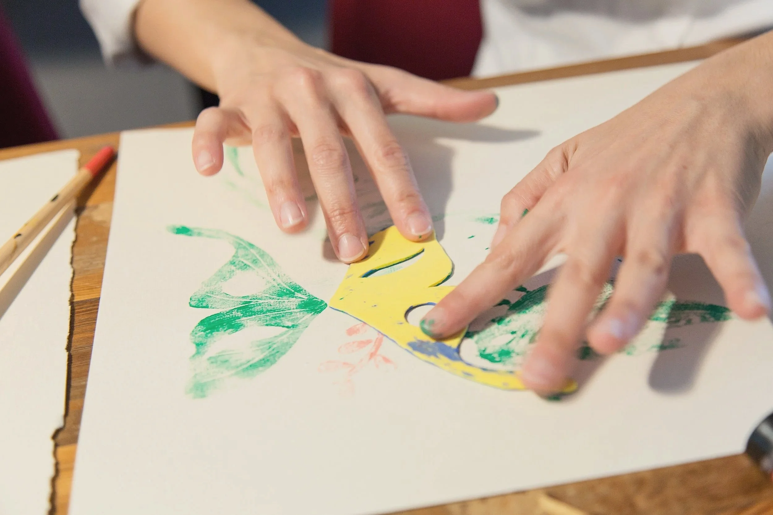 Hands creating artwork with green, yellow, and blue paints on paper.