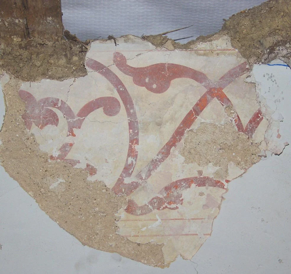 Old, damaged wall with a faded decorative mural in red and pink hues, partially covered with dirt and crumbling plaster.
