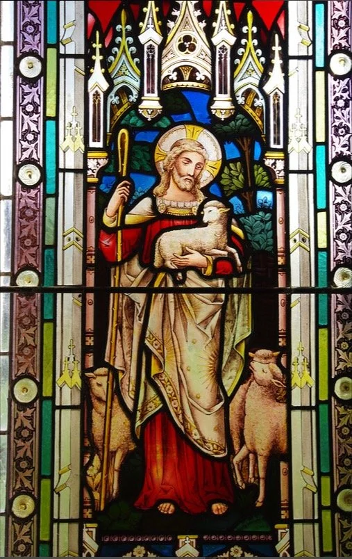 Stained glass window depicting Jesus Christ holding a lamb, surrounded by a decorative gothic architectural frame.