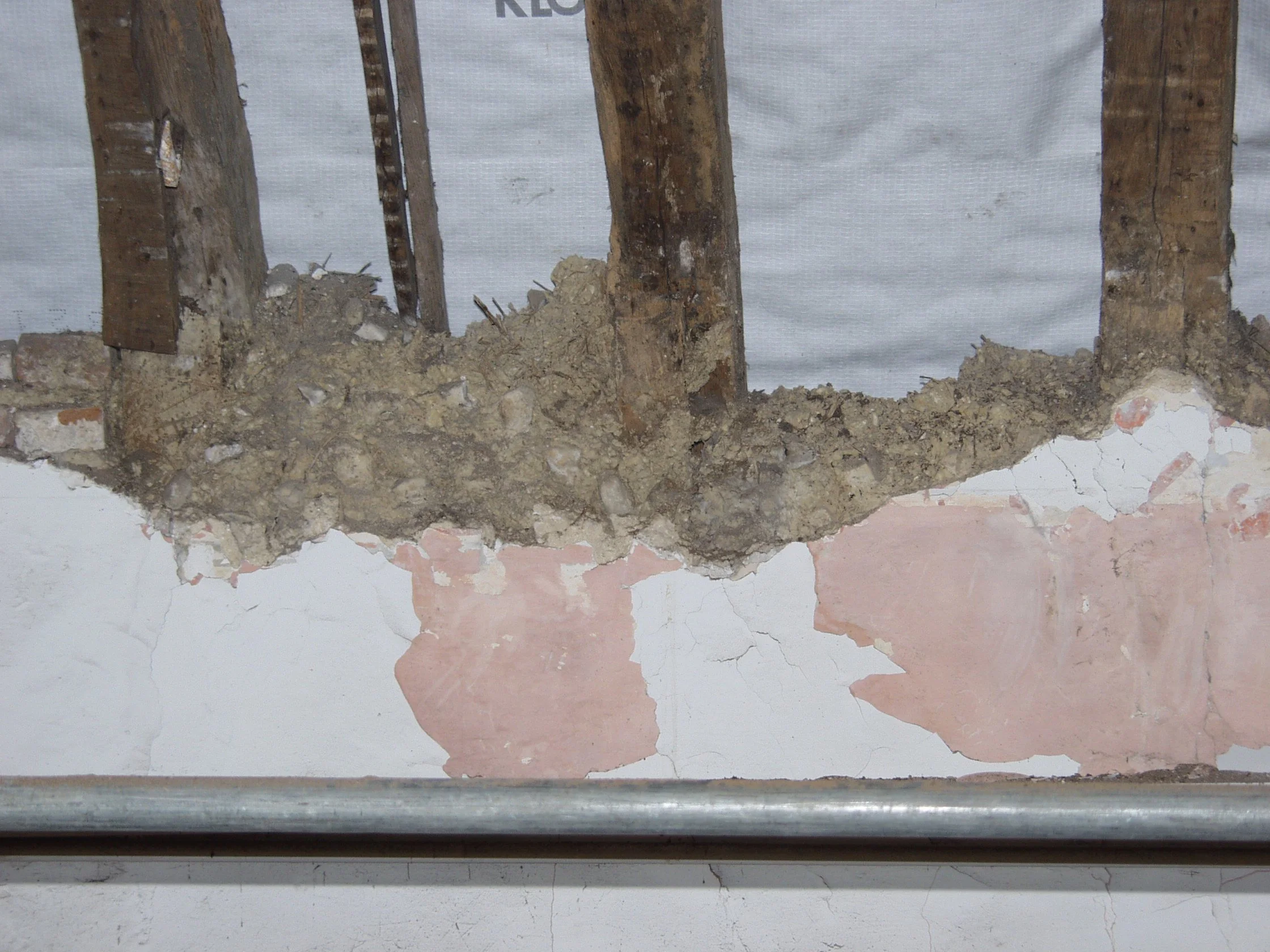 Close-up of a section of a wall with exposed wooden studs, damaged drywall, and visible insulation, with a metal pipe running horizontally in the foreground.