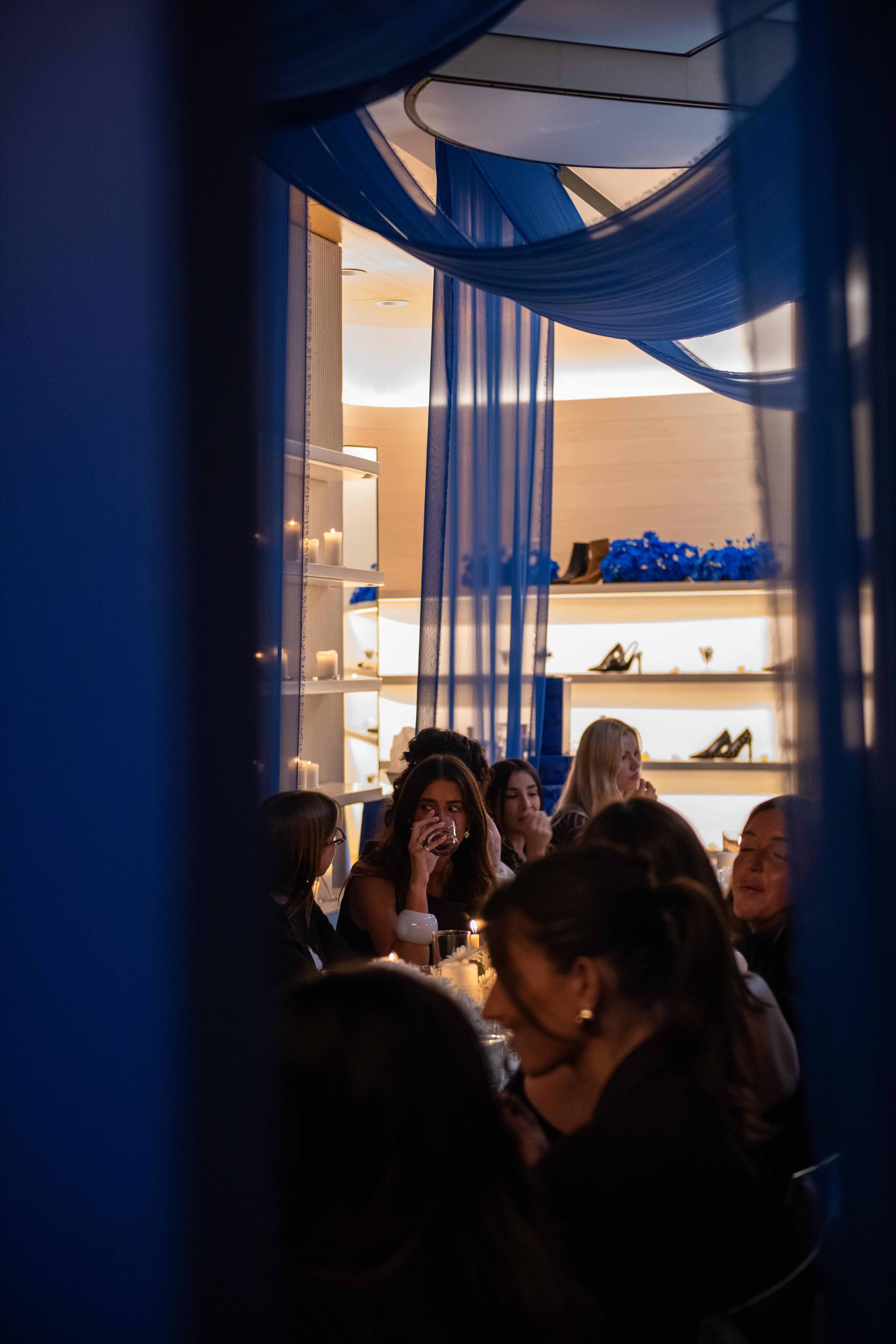 Women celebrating at a dinner party in a modern venue with blue curtains and shelves displaying shoes and candles.
