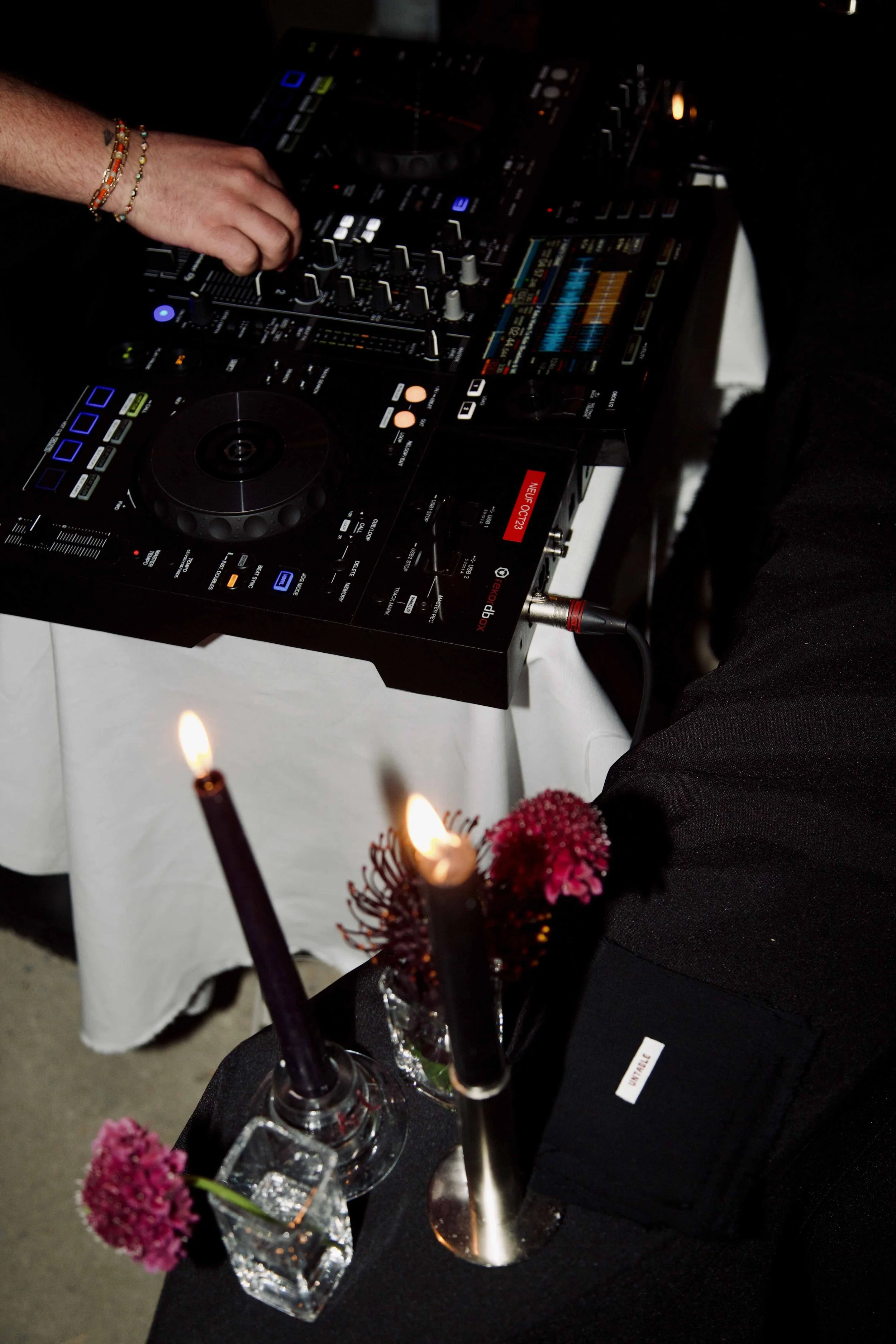 A DJ is mixing music with professional DJ equipment, including a mixer and turntables, on a table covered with a white cloth. There are three lit candles and pink flowers in vases nearby.