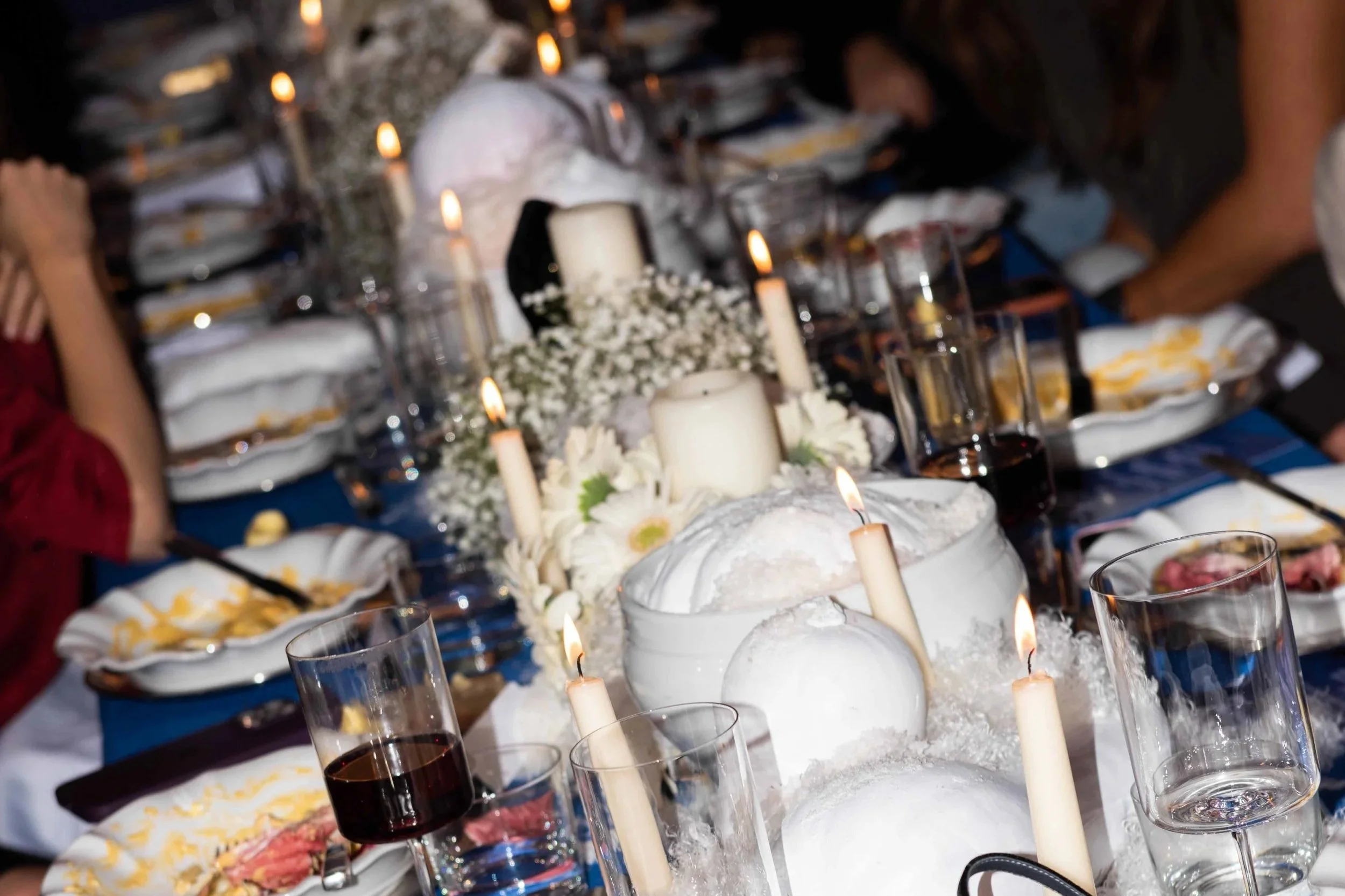 A decorated dining table set for a formal event with white candles, floral centerpieces, glasses of wine, and place settings, with some people seated around it.