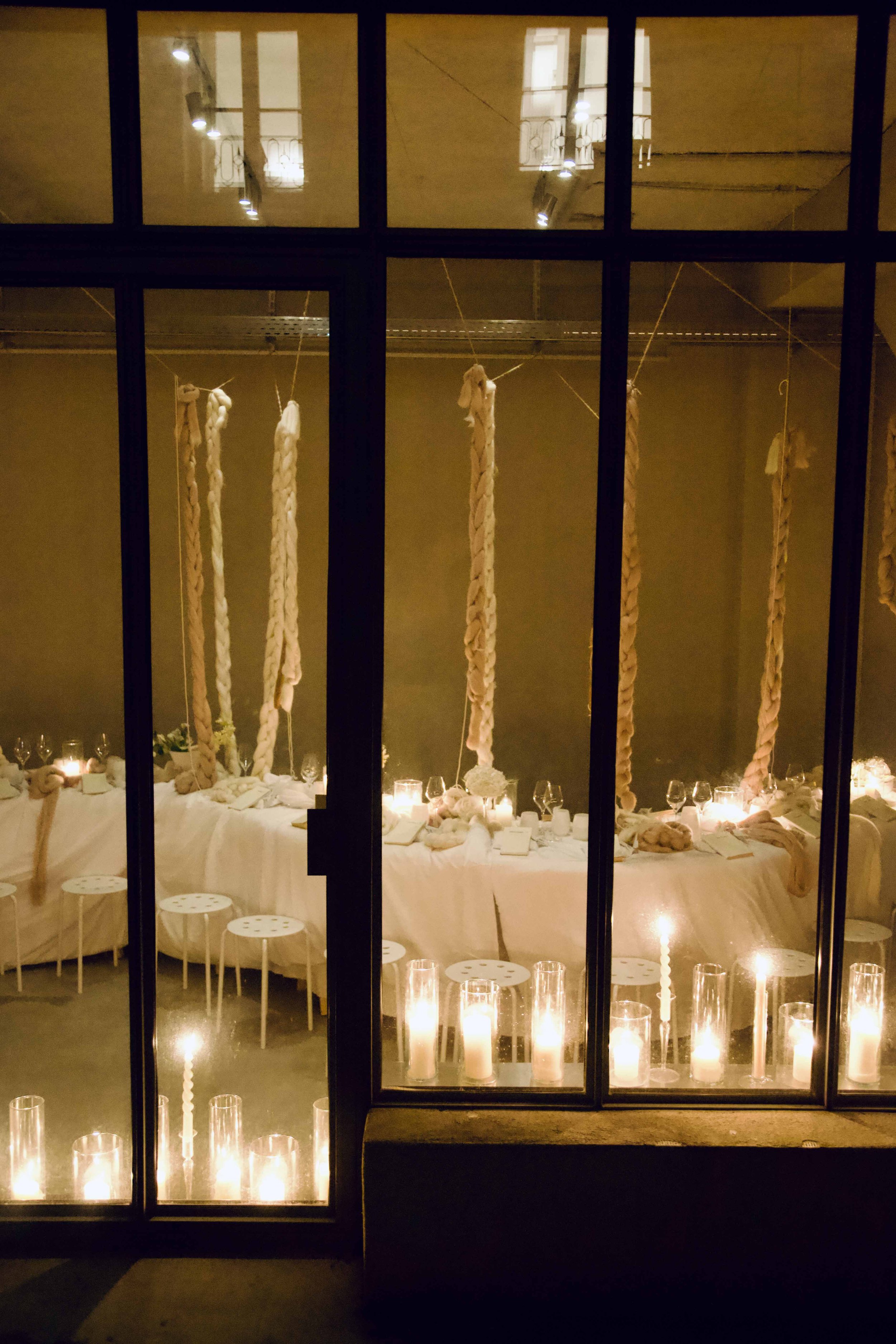A dining area seen through glass doors, decorated with large twisted fabric hanging from the ceiling, and luminary candles on the table and on the window sill, creating a warm, intimate ambiance.