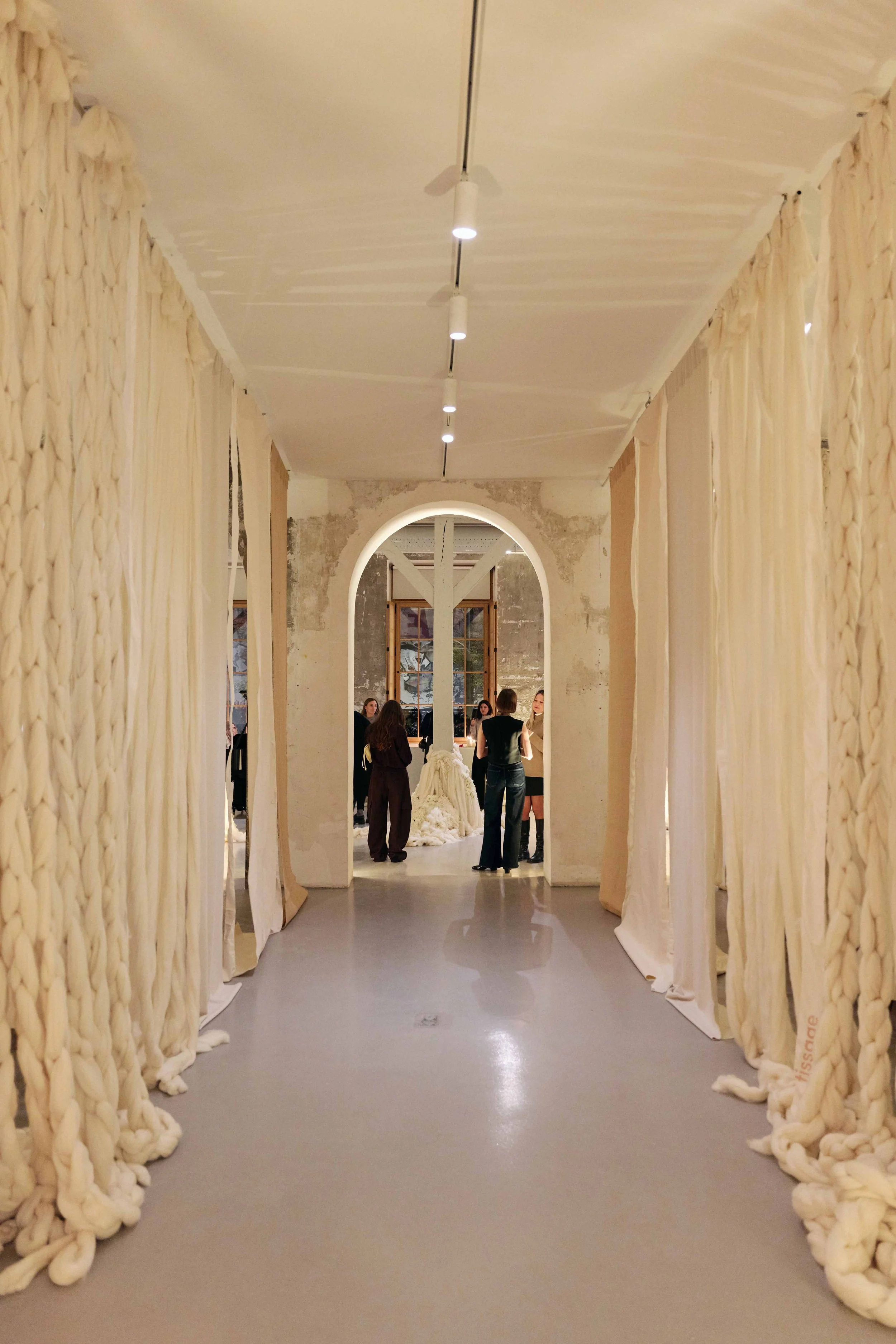 A hallway leading to a room with people gathered, decorated with knotted fabric curtains on the sides and a large arched opening in the background.