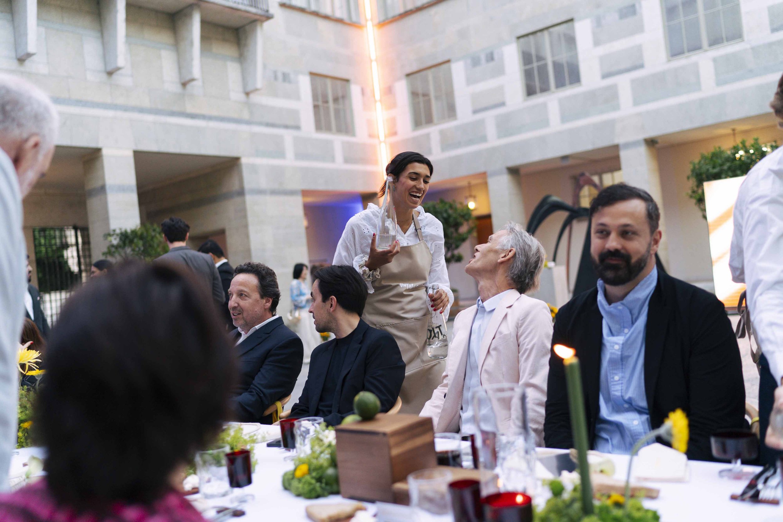People sitting at a banquet table in an indoor venue, a woman in an apron serving drinks, others engaging in conversation and smiling.