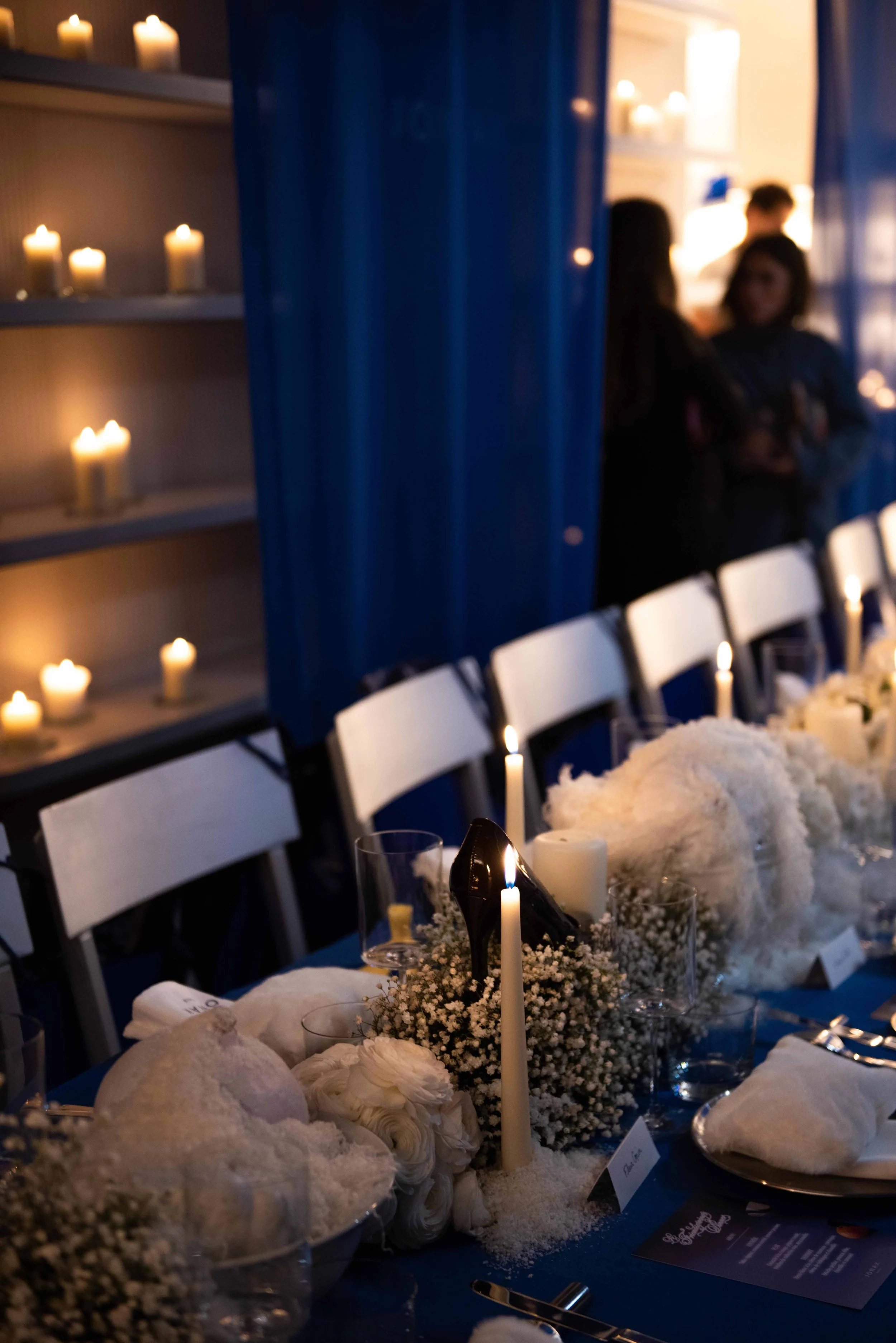 A wedding reception table decorated with white flowers, candles, and black high heel shoes as a centerpiece, with a background of blue curtains and candles on shelves.