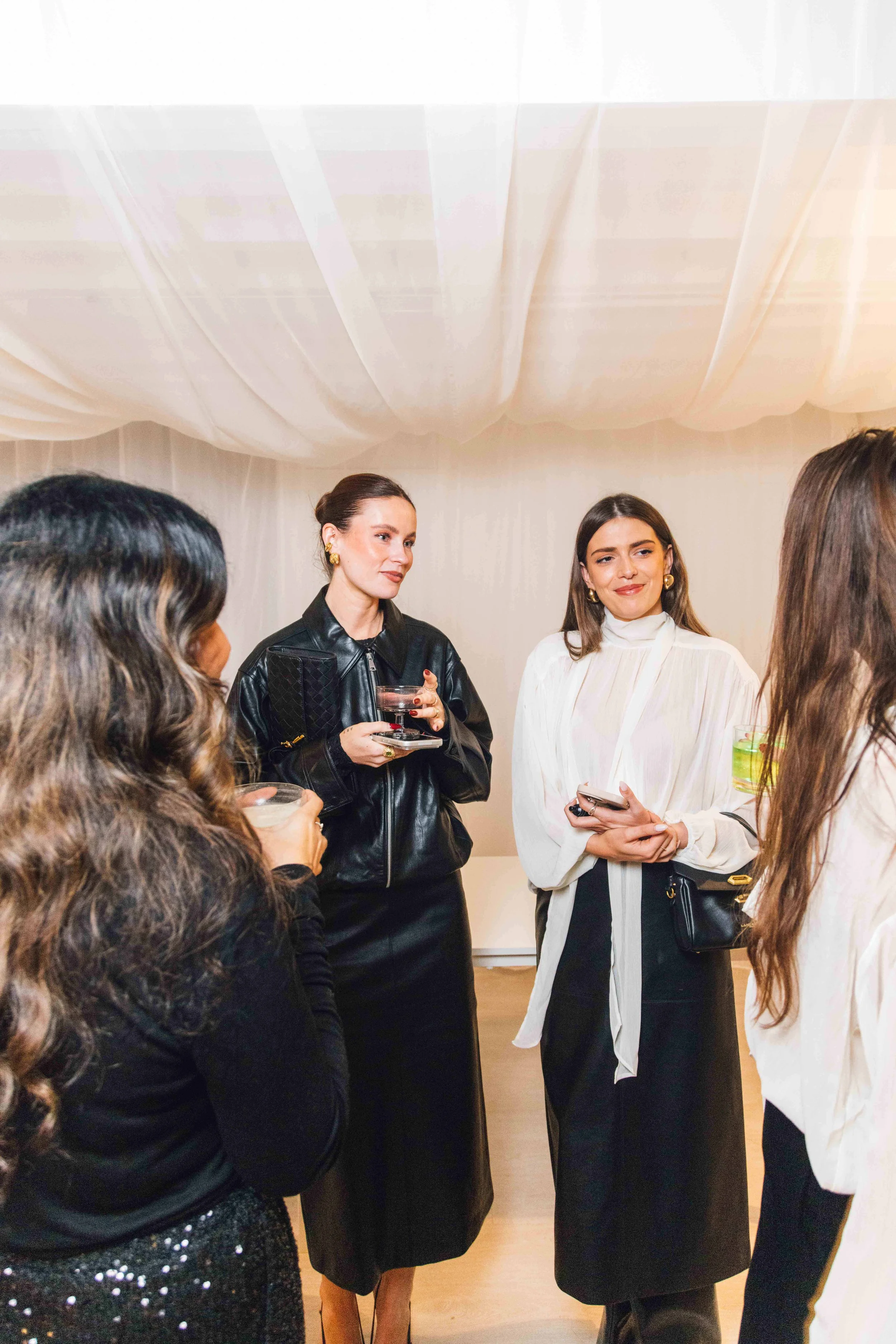 Group of women at a social gathering, holding drinks, in a room with white draped ceiling, dressed in stylish outfits.