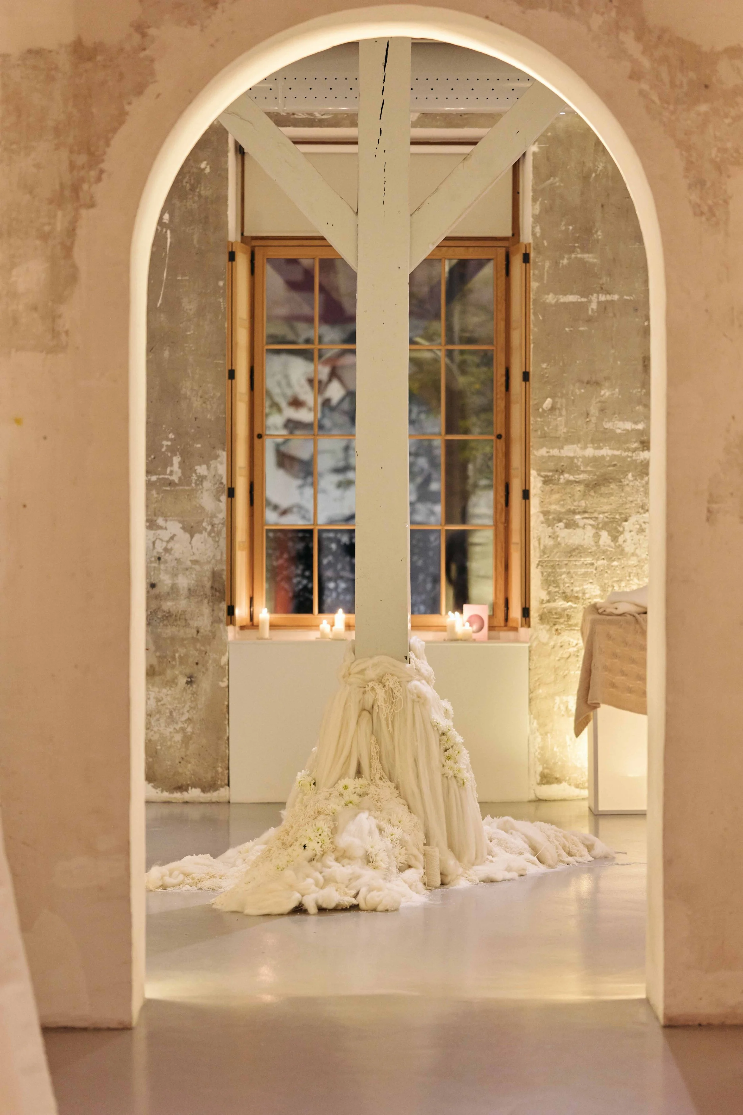 A white fabric draped cross embedded with flowers, displayed in front of a window with candles on the windowsill, seen through a rounded archway in a room with exposed stone walls.