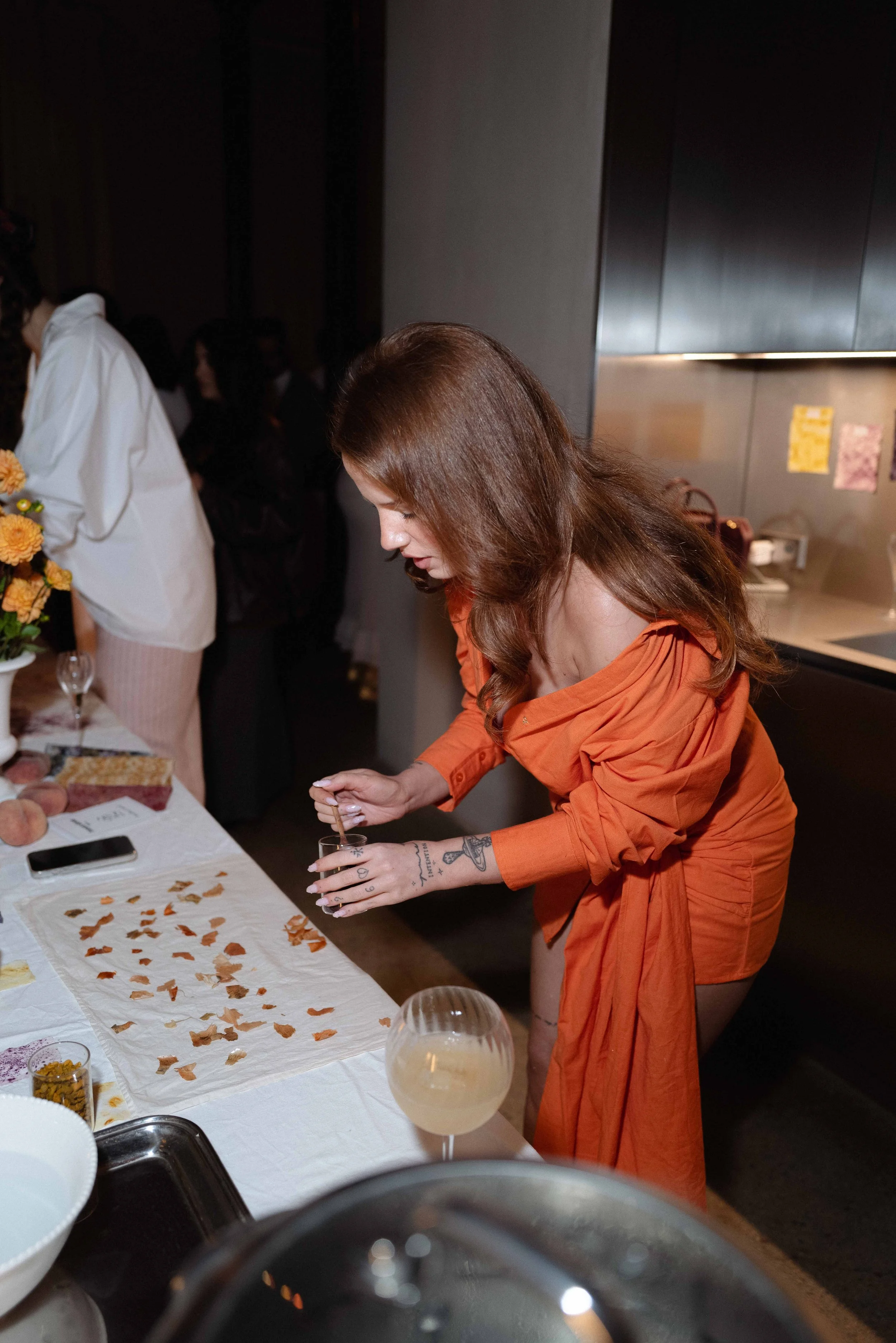 A woman with long brown hair, wearing an orange dress, is preparing a drink at a social gathering. She is standing at a table with food and flowers, pouring a liquid into a glass. The background shows other people and a kitchen area.