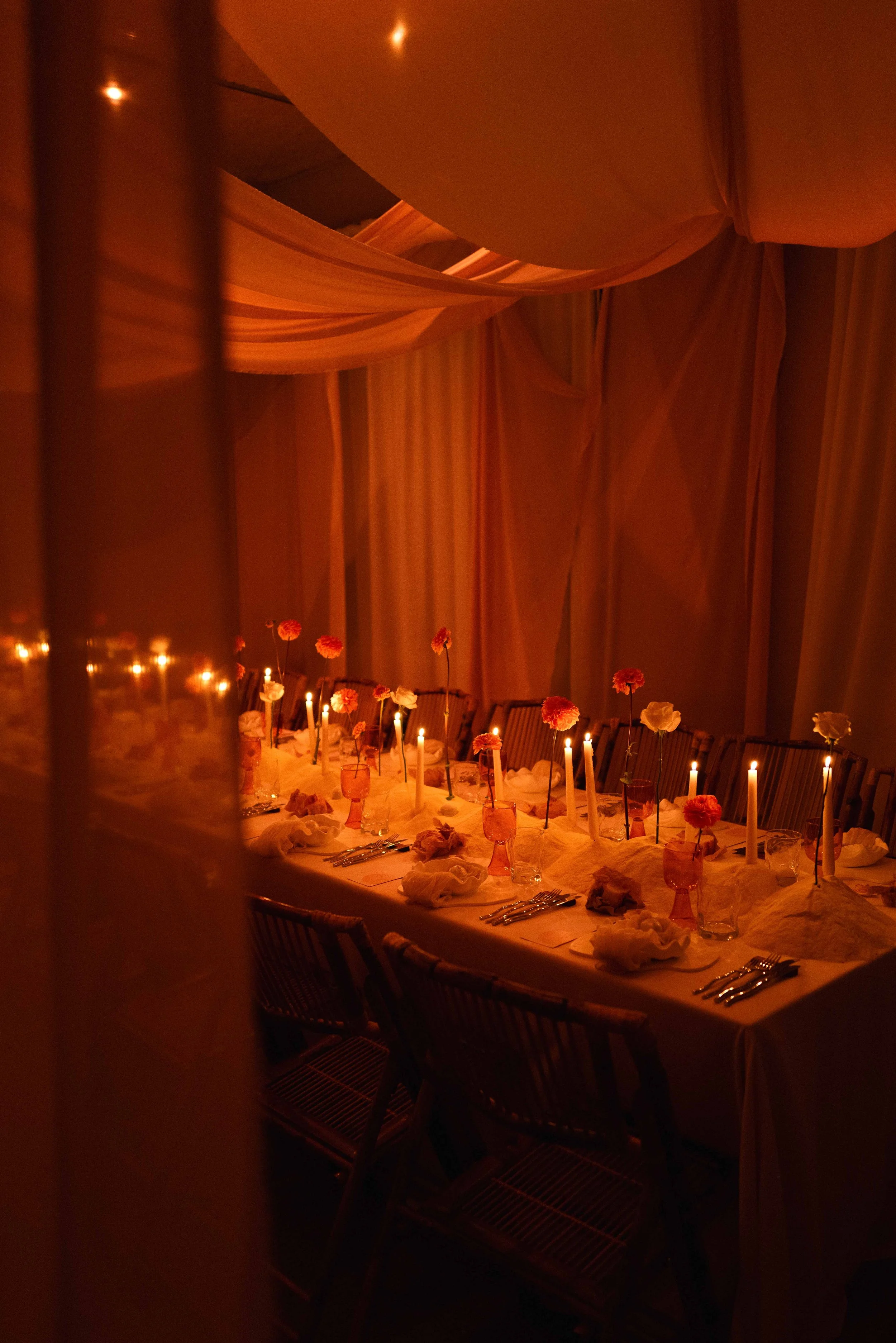 A long dining table set for a special occasion with lit candles, flowers in pink and white, glassware, and folded napkins, under warm orange lighting with draped fabric overhead.