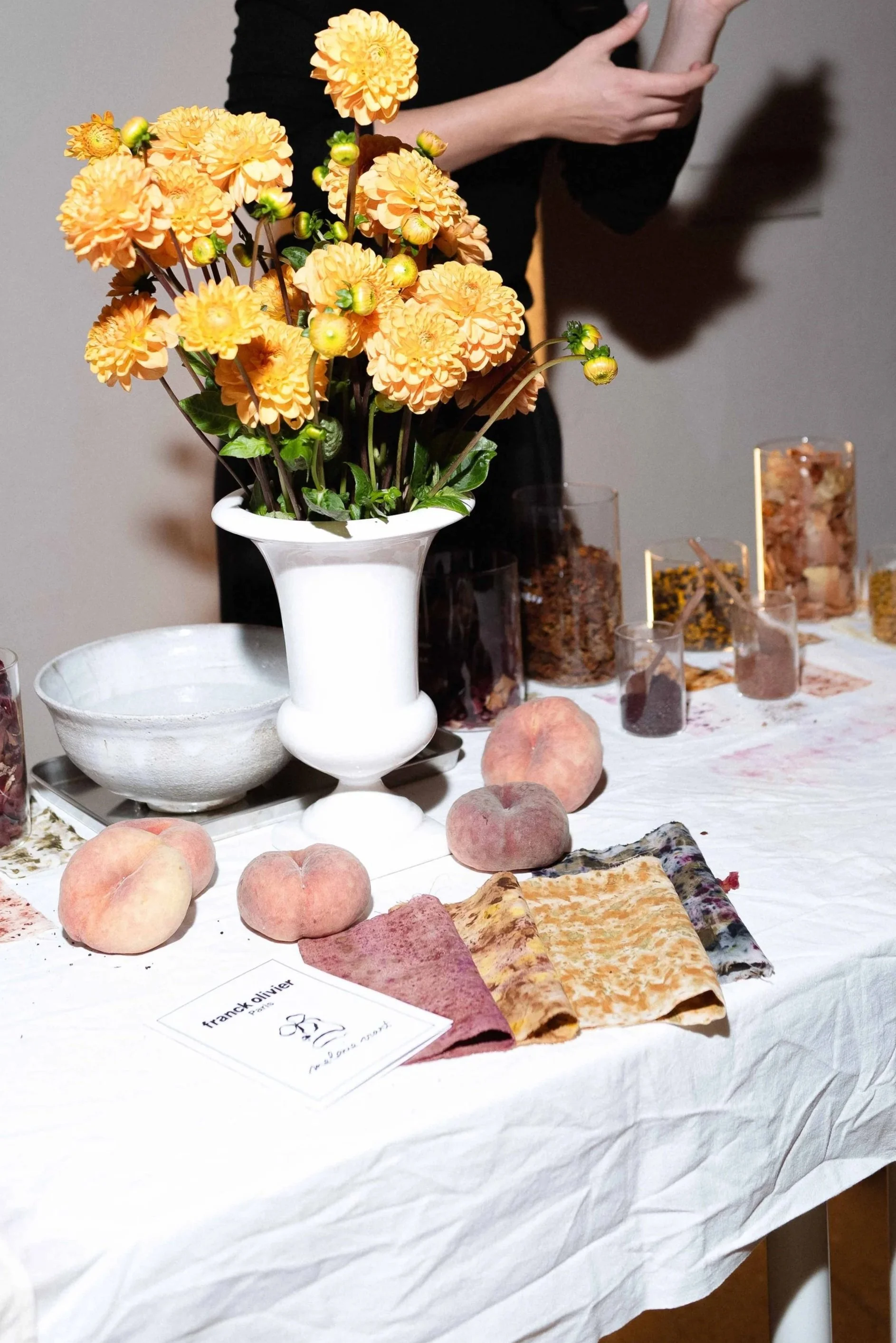 A floral display with a large bouquet of orange flowers in a white vase, peaches on the table, patterned fabric samples, and glass containers with herbs or spices. A person is partially visible in the background, reaching across the table.