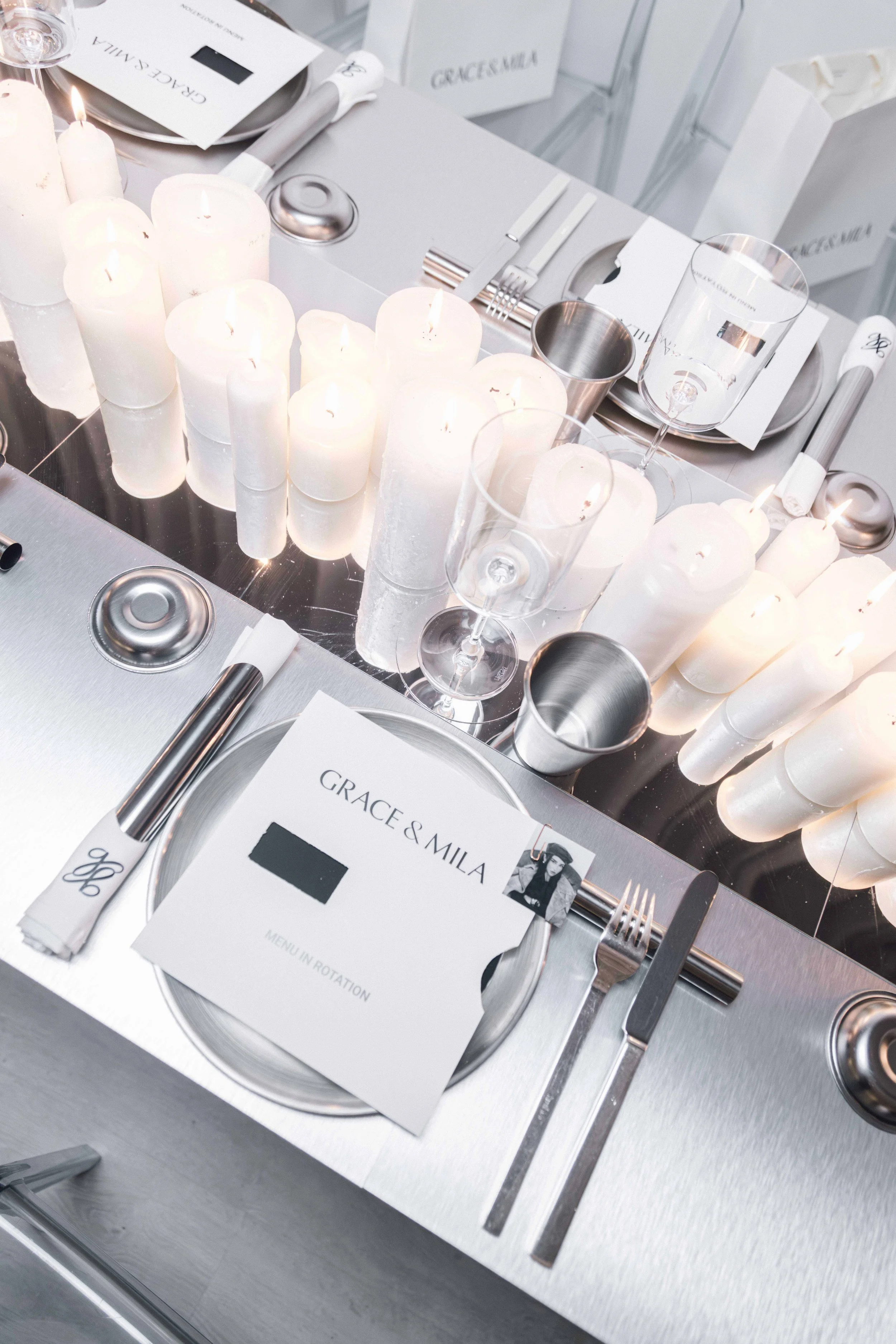 Wedding table setting with white candles, metallic dinnerware, wine glasses, a menu card labeled 'Grace & Mila', and silver and white decor.