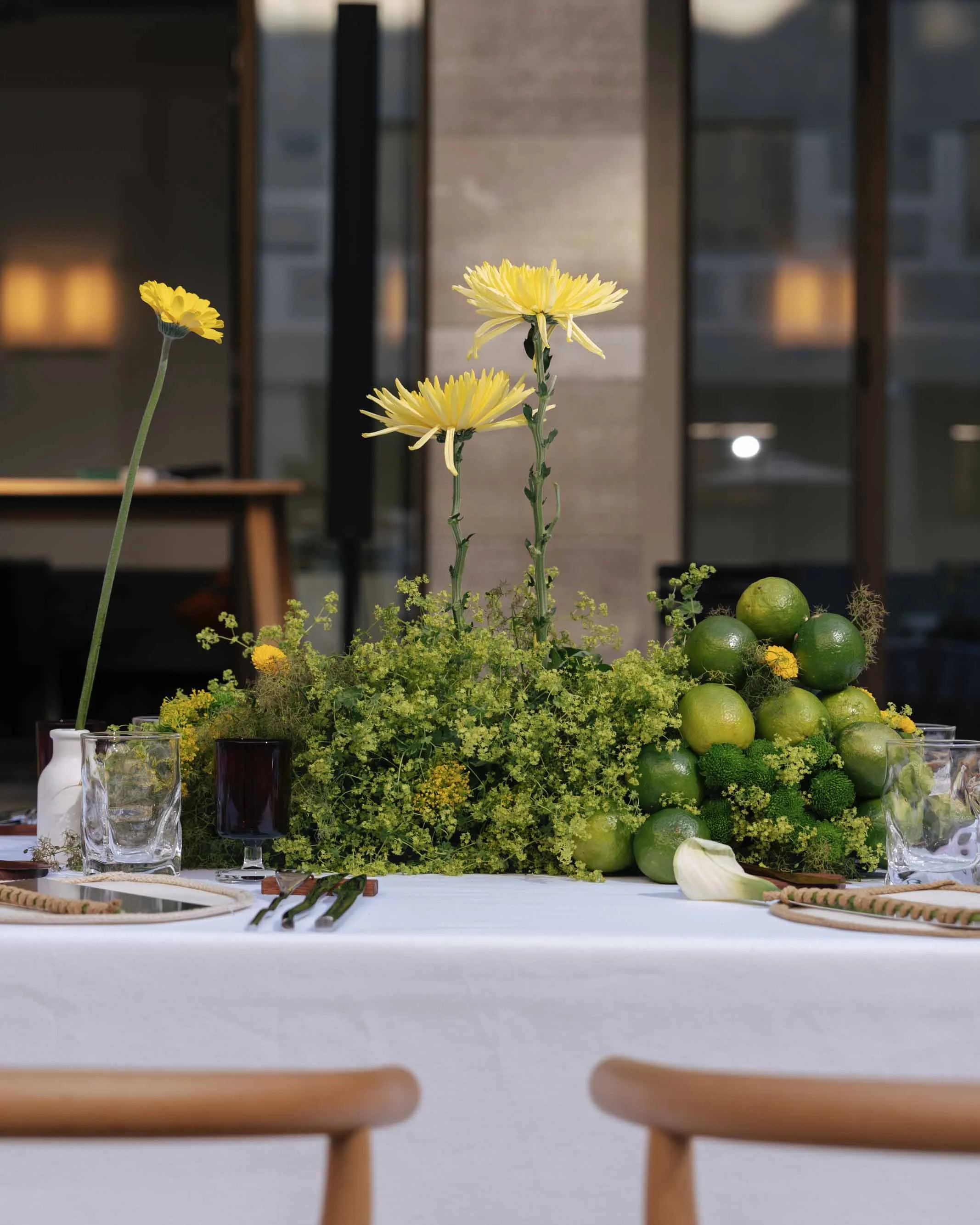 Elegant table centerpiece with yellow flowers, green limes, and lush greenery on a white tablecloth.