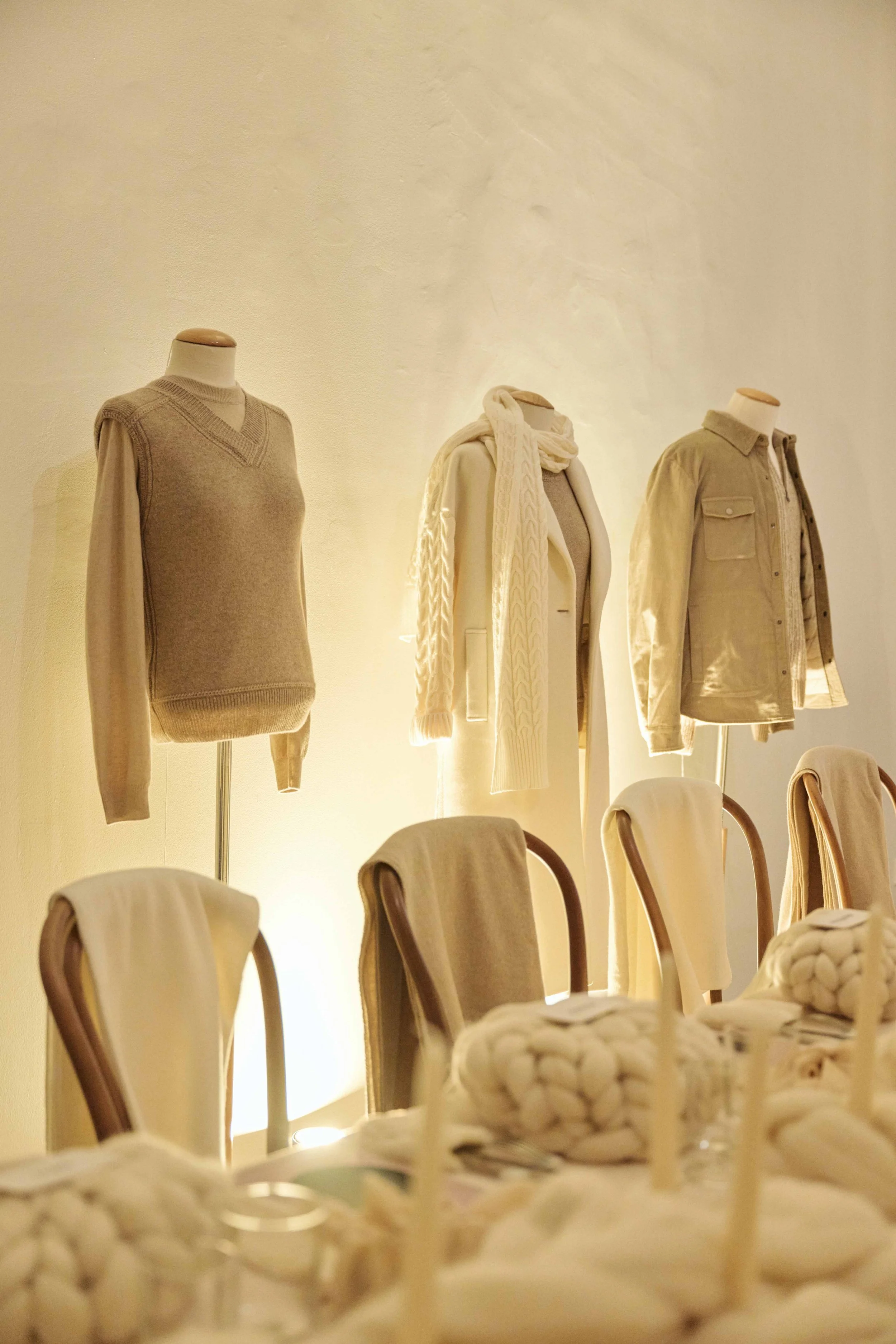 Men's and women's clothing mannequins dressed in neutral-colored sweaters, shirts, and a coat in a softly lit setting, with chairs and decorative items in the foreground.