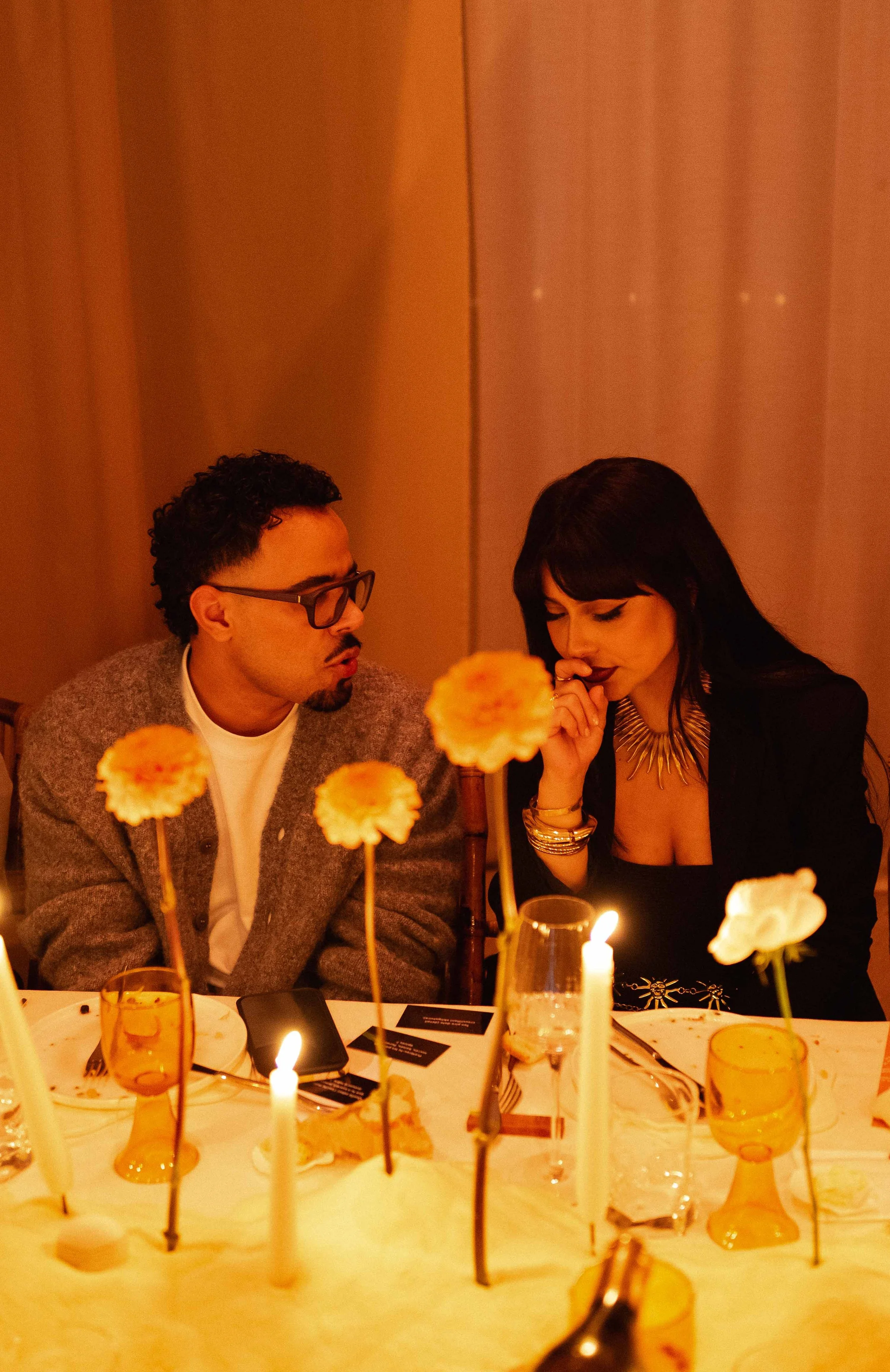 Two people sitting at a dinner table with lit candles and decorated with flowers, engaged in conversation.