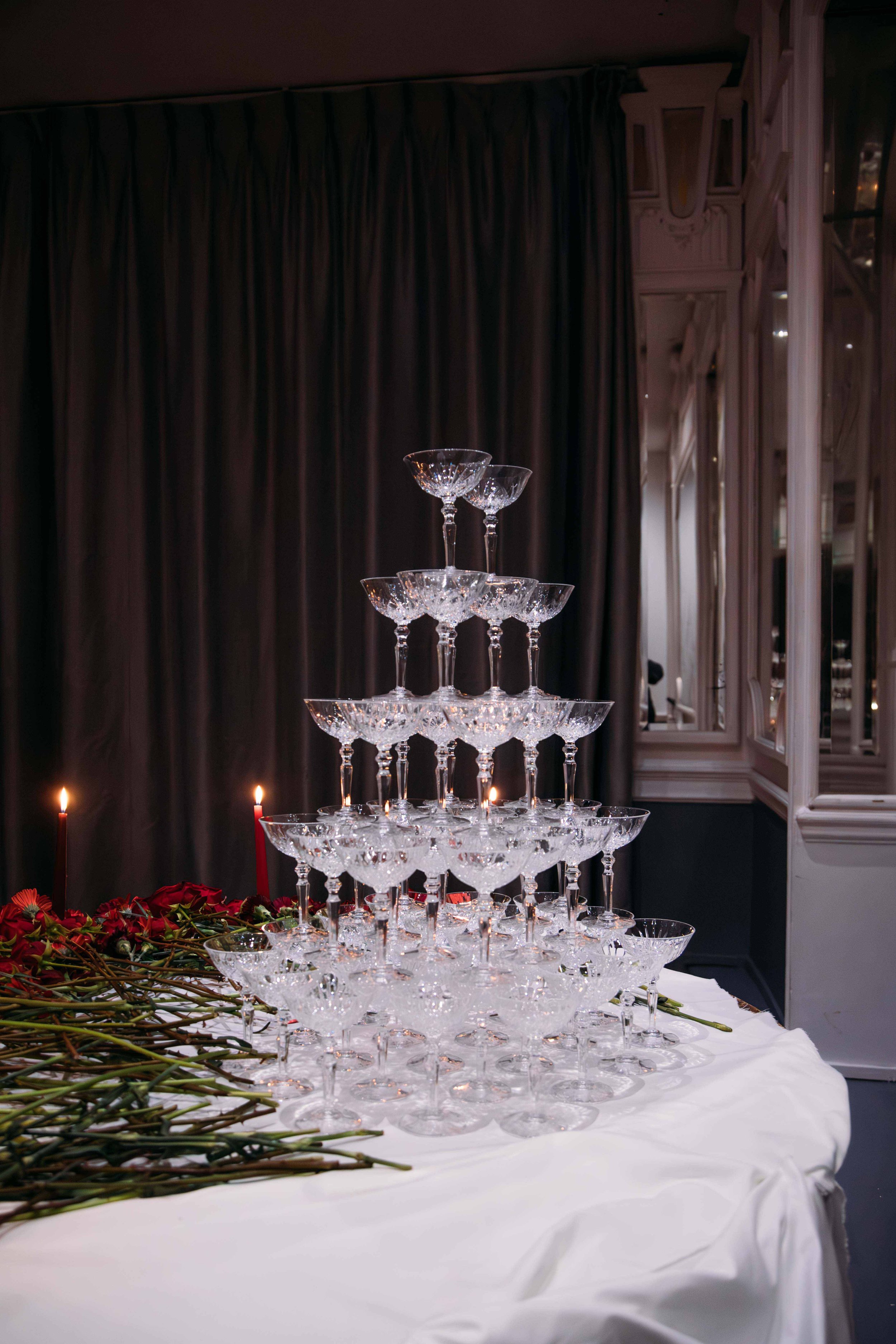 A pyramid of empty champagne glasses on a white table with red roses and lit candles, against a dark curtain and ornate wall mirrors.