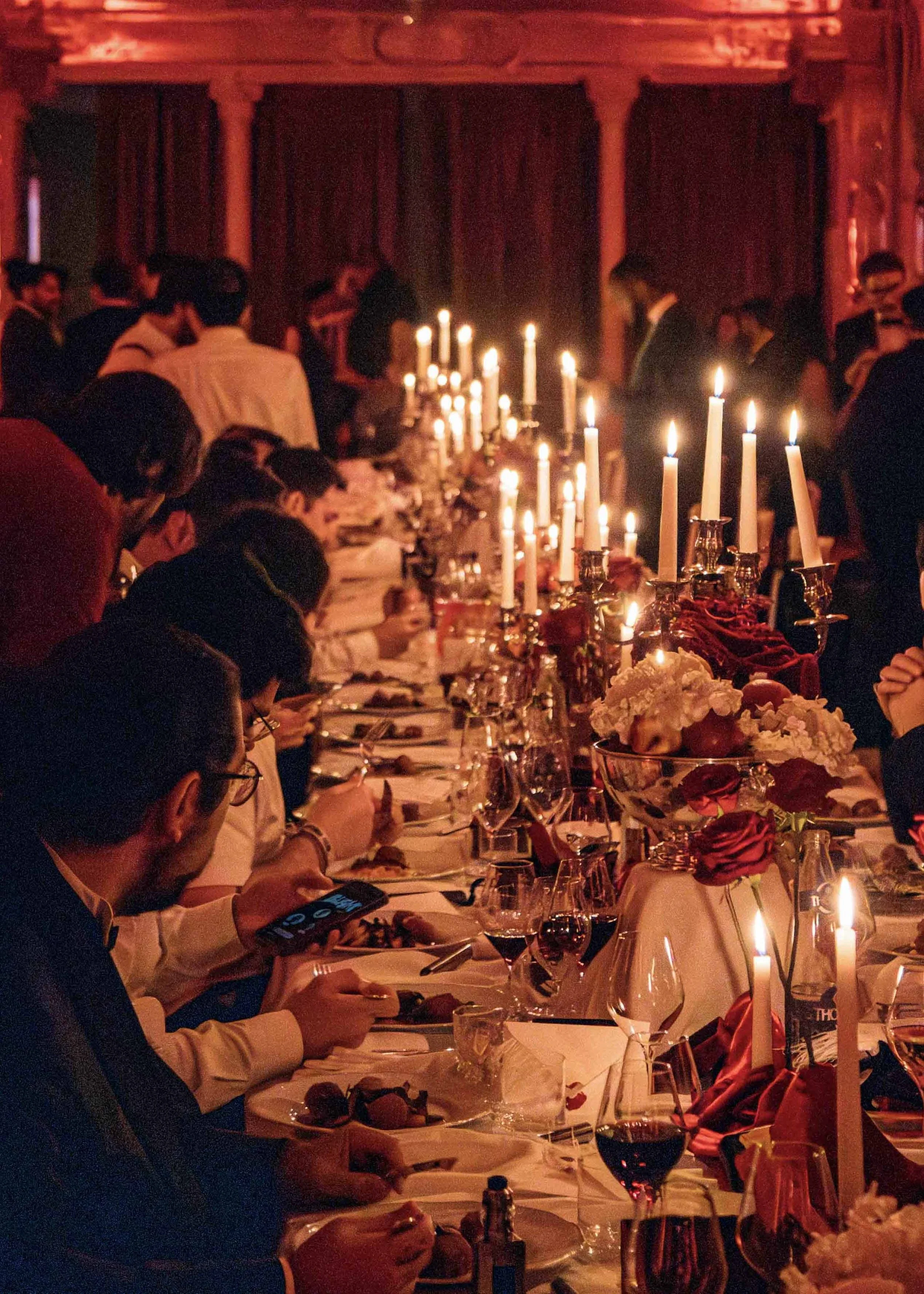 A large dimly lit banquet room with a long table decorated with candles, flowers, and elegant tableware. People are seated on both sides of the table, dining and socializing.