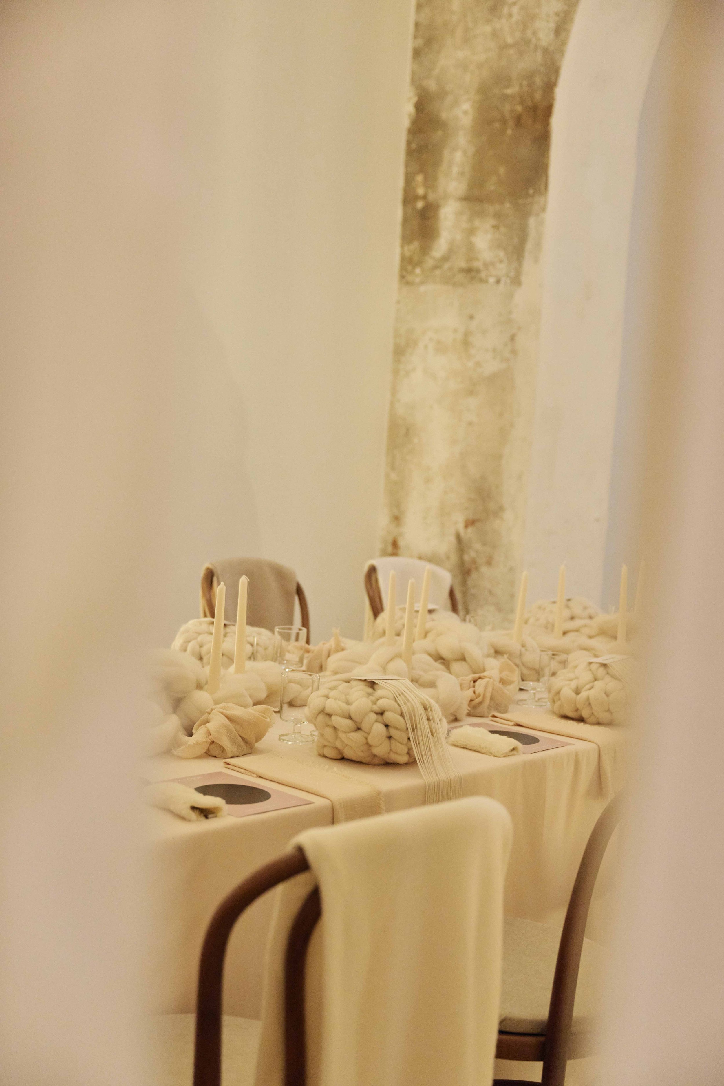 Dining table set with beige tablecloth, industrial-looking chunky wool centerpieces, white candles, and minimalist tableware in a warmly lit room with exposed stone wall and chairs around the table.