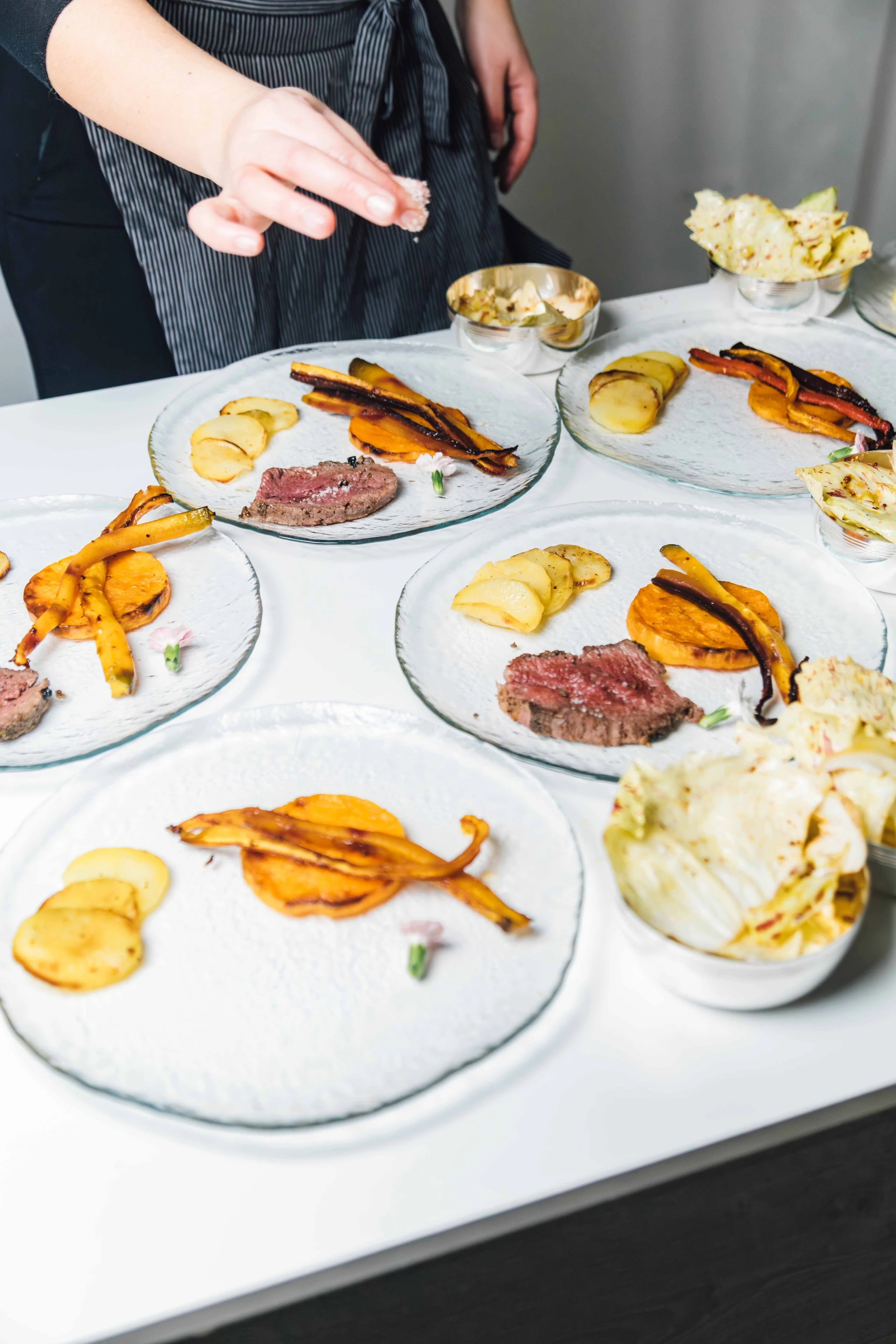 Plates with sliced beef, roasted carrots, roasted potatoes, and grilled red and yellow bell peppers on a table. Someone's hand is sprinkling salt on one of the dishes.