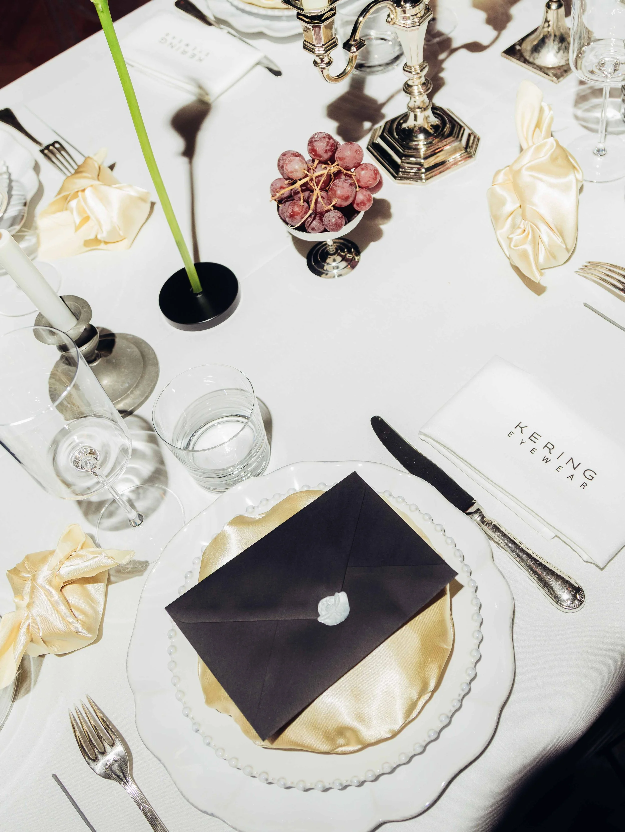 A formal dining table set with champagne flutes, water glasses, silverware, beige satin napkins, a black envelope with a white wax seal on a gold satin placemat, a small bowl of red grapes, a silver candlestick, and a silver candelabra on a white tablecloth.