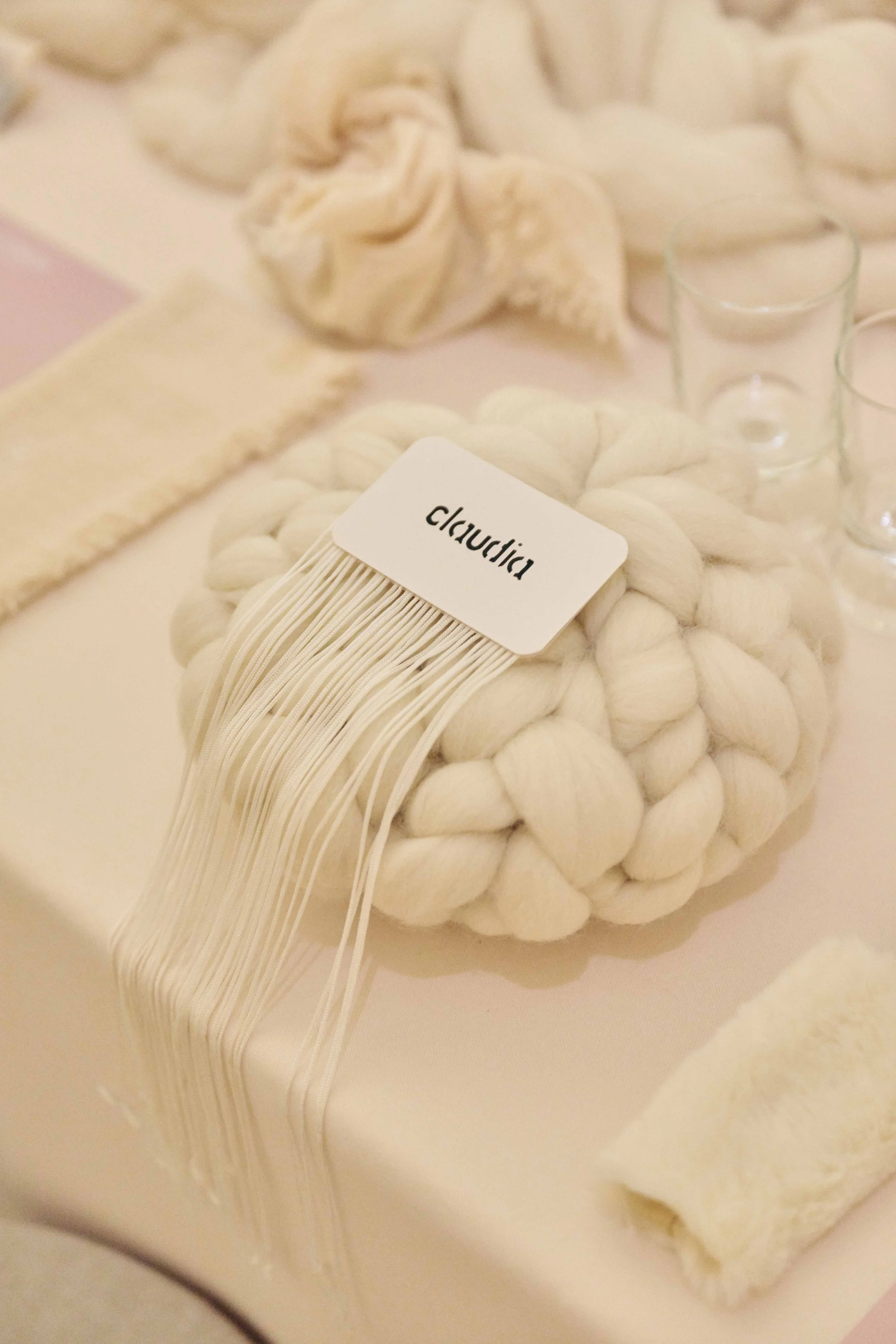 A bundle of white wool yarn labeled 'claudià,' placed on a soft pink table with glass votive candle holders and fabric displayed nearby.