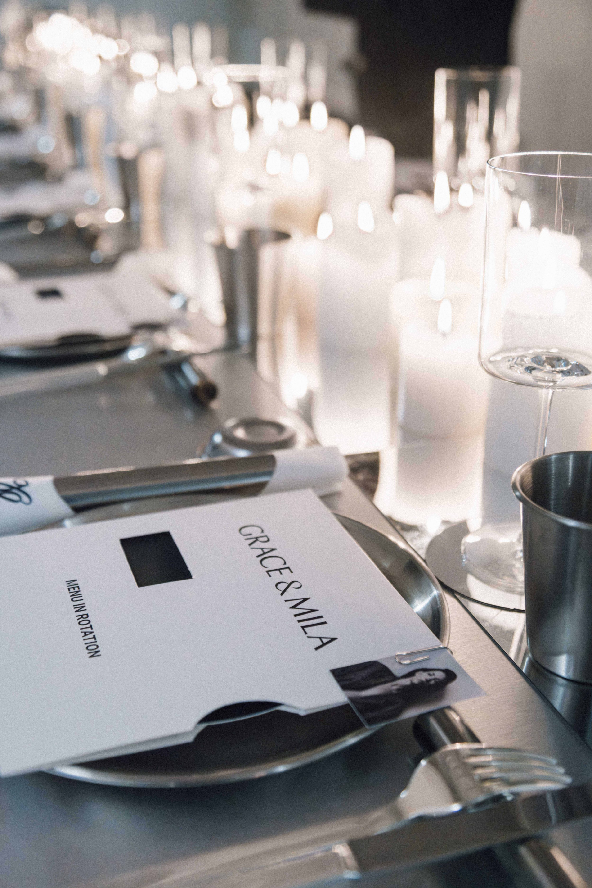 A wedding place setting with a table card that reads 'Grace & Mila,' a black and white photo of a woman, a pen, clear glass candlesticks with white candles, empty wine glasses, and silver utensils on a silver tray.