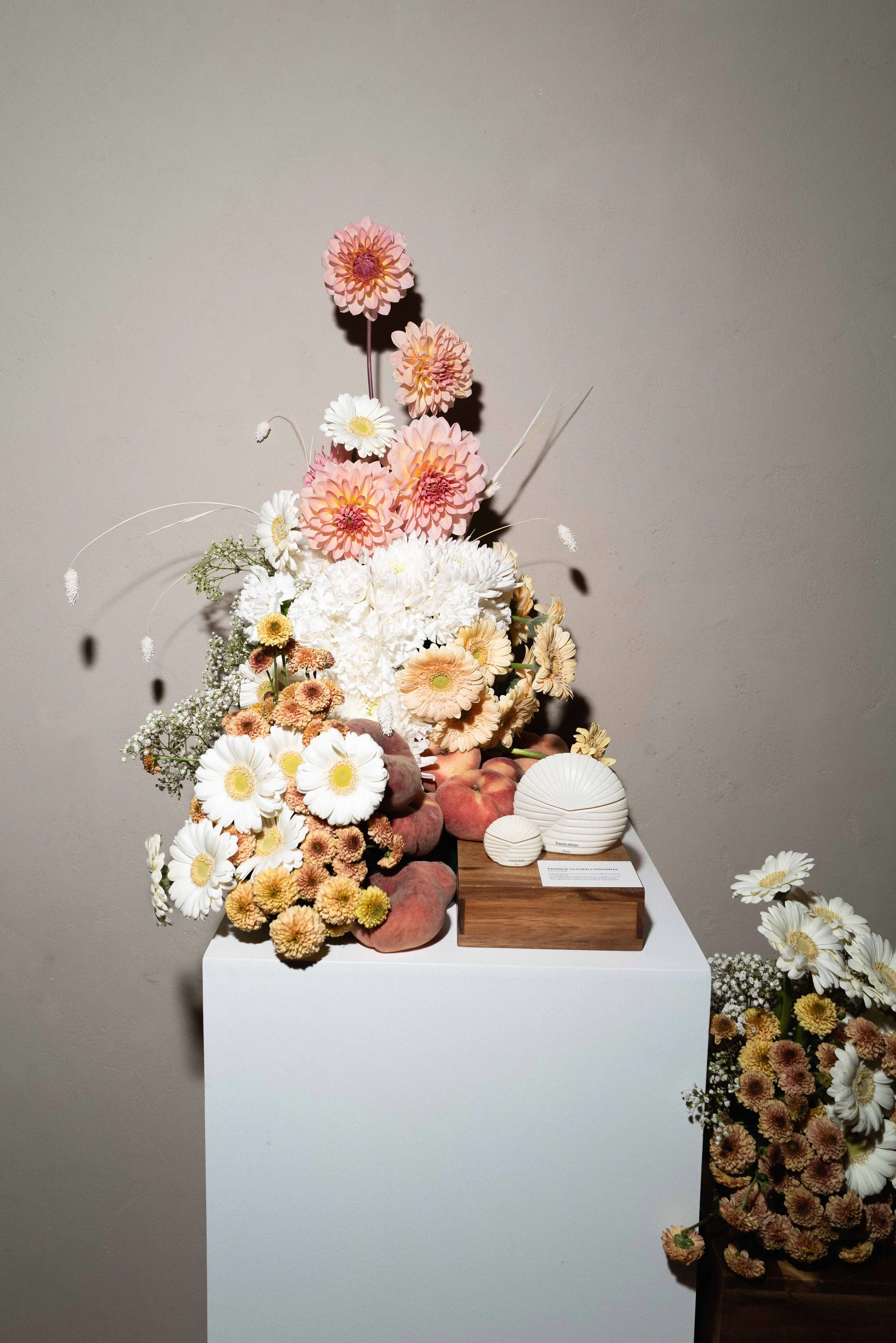 Display of white, pink, and peach-colored flowers arranged with peaches and decorative white ceramic objects on a white pedestal.