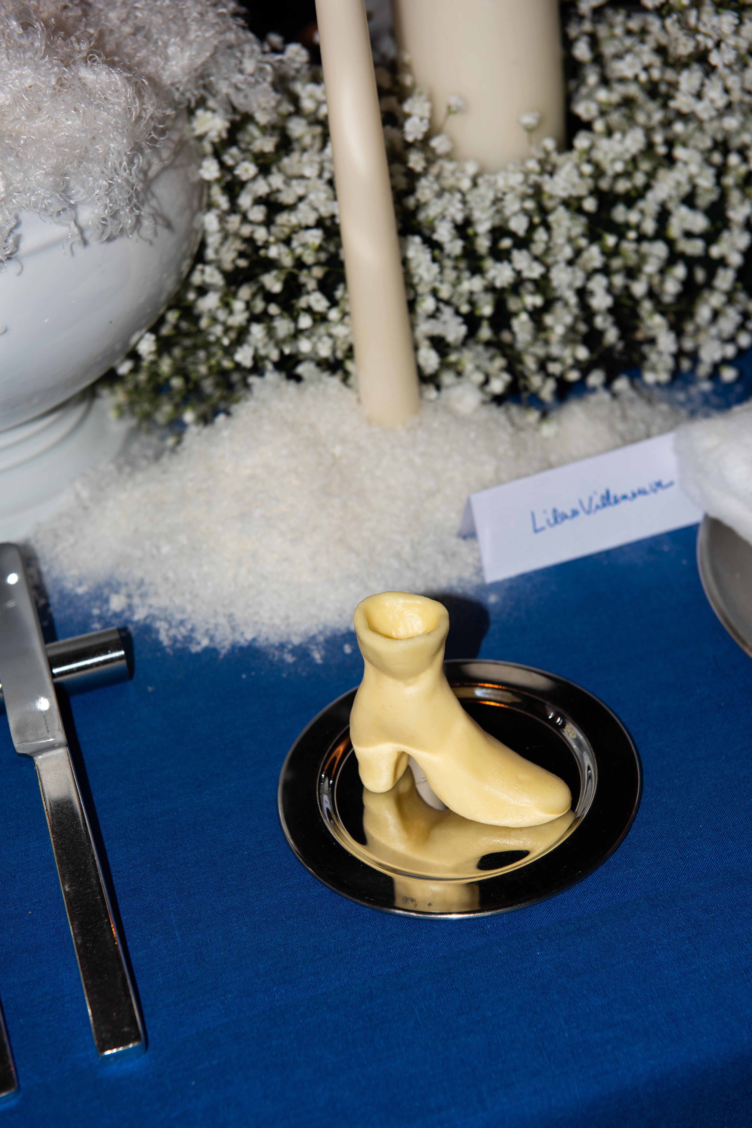 White chocolate shoe cake on a silver plate placed on a blue tablecloth. In the background, a table with white candles surrounded by white flowers and a name card with 'Lina Vulgione' written.