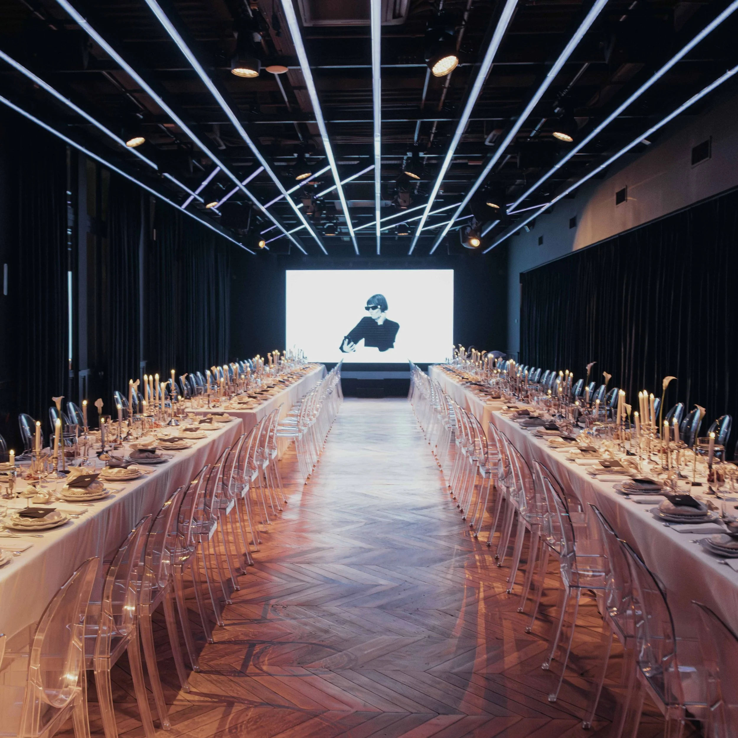 Elegant banquet hall with long tables set for a formal dinner, featuring candles and glassware, with a large bright screen at the front displaying a black-and-white image of a woman wearing sunglasses.