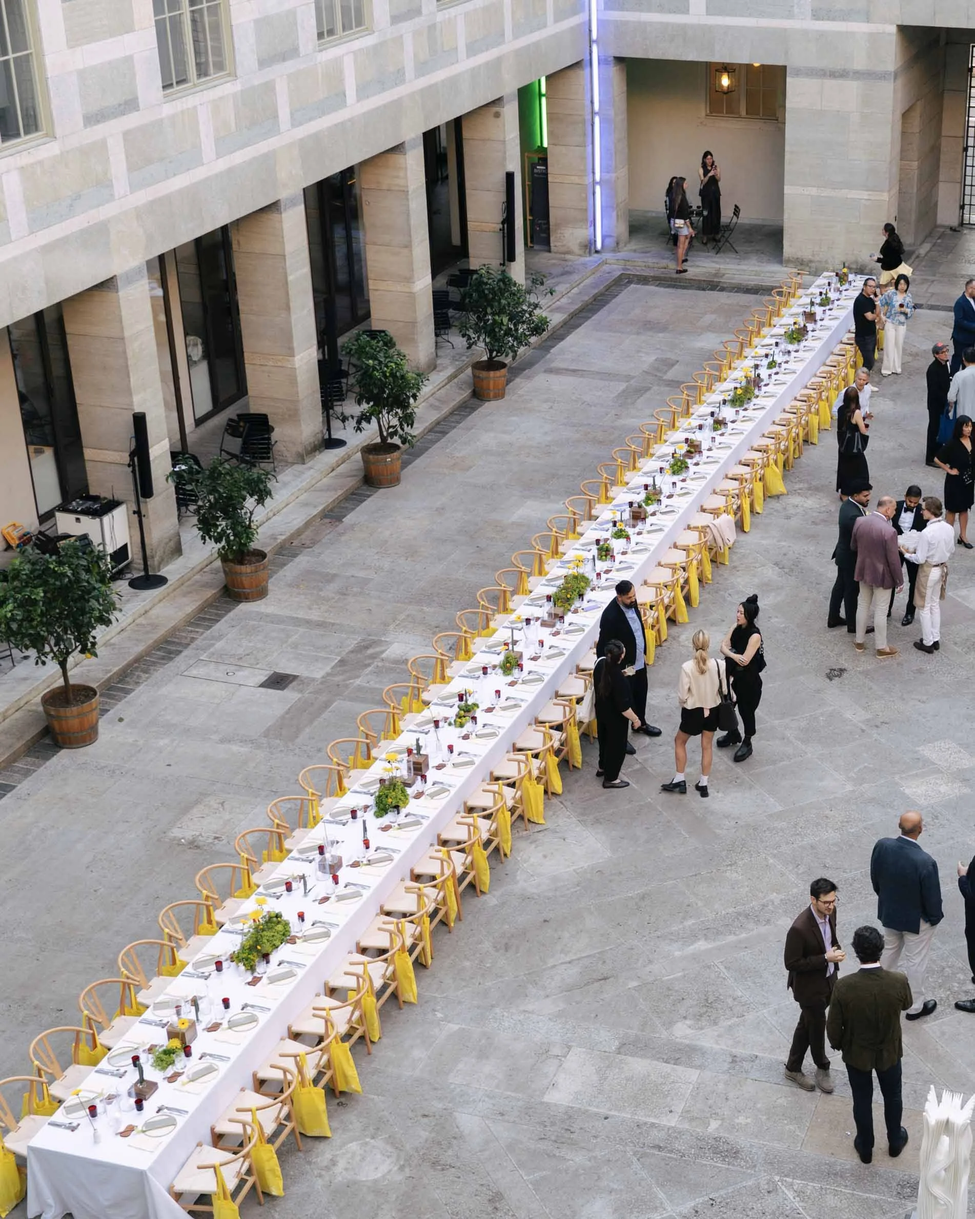 Long banquet table set for a formal event with yellow chairs, floral centerpieces, wine glasses, and place settings, located in an indoor courtyard with potted plants and several guests mingling nearby.