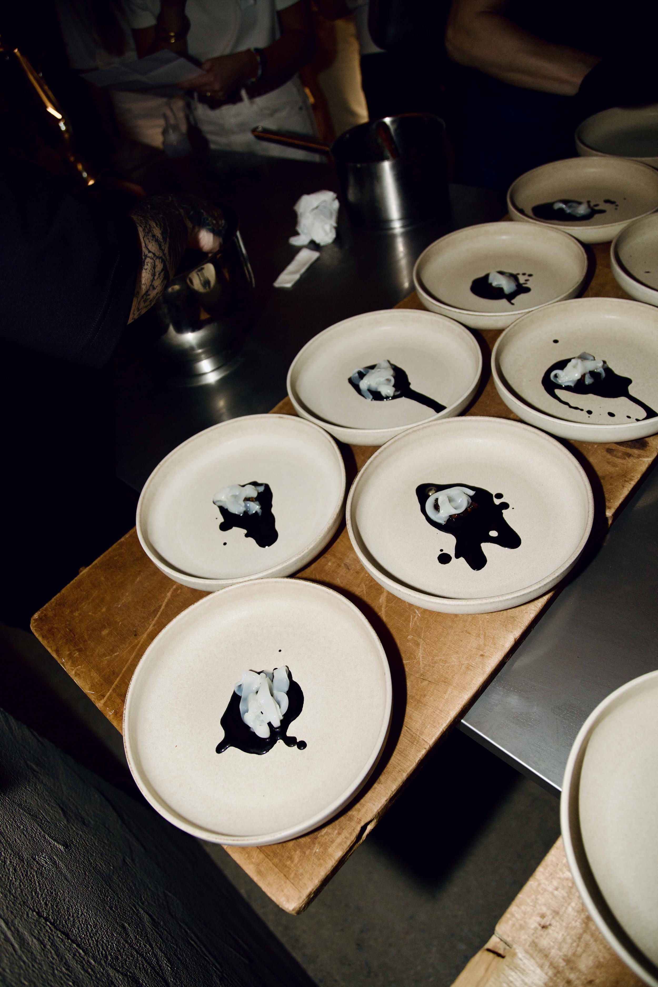 Several empty beige plates with white dollops of black sauce on a wooden table, with some people standing in the background.