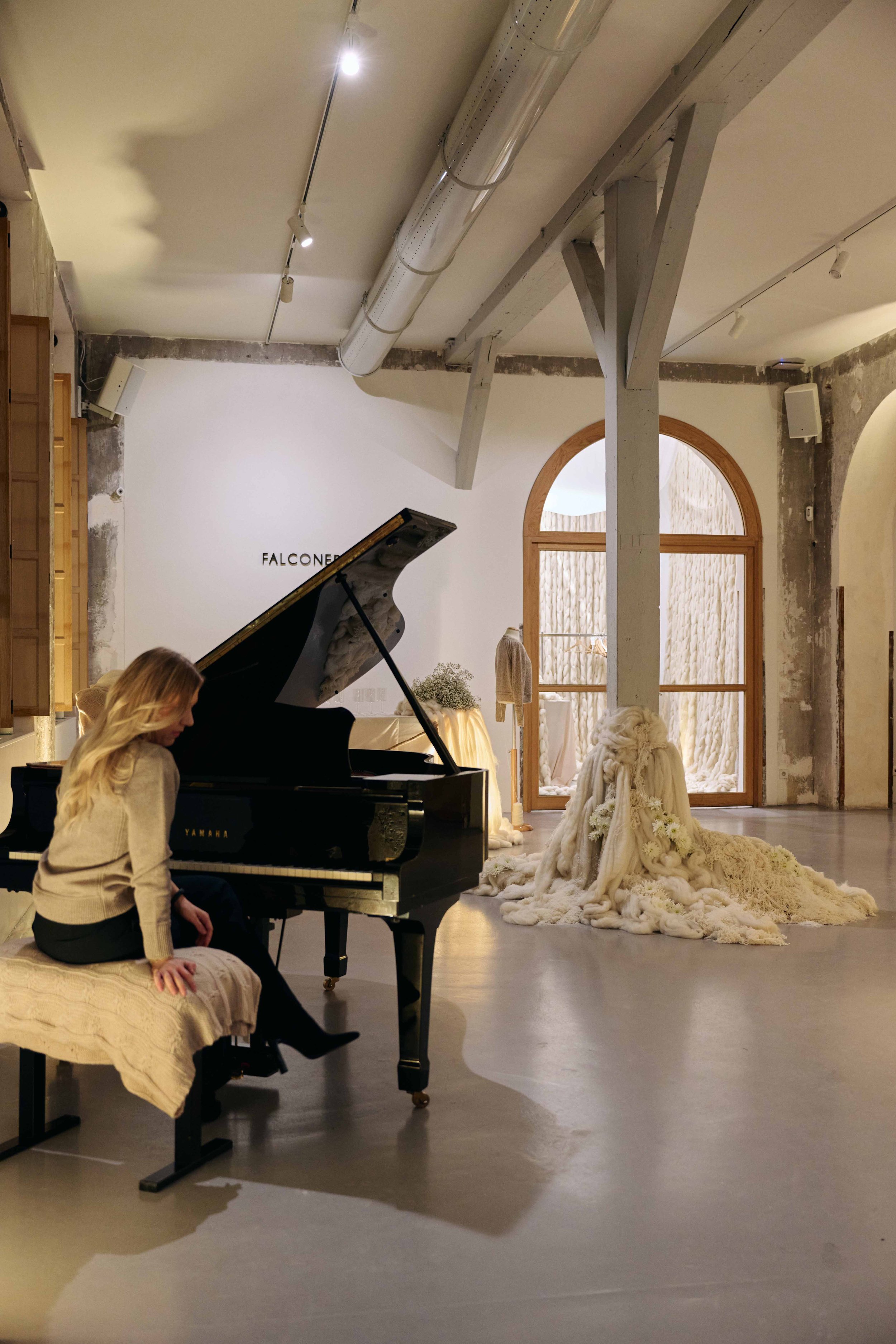 A woman sitting at a black piano in an art gallery or studio, with a sculpture draped in white fabric resembling a person, and a large arched window in the background.