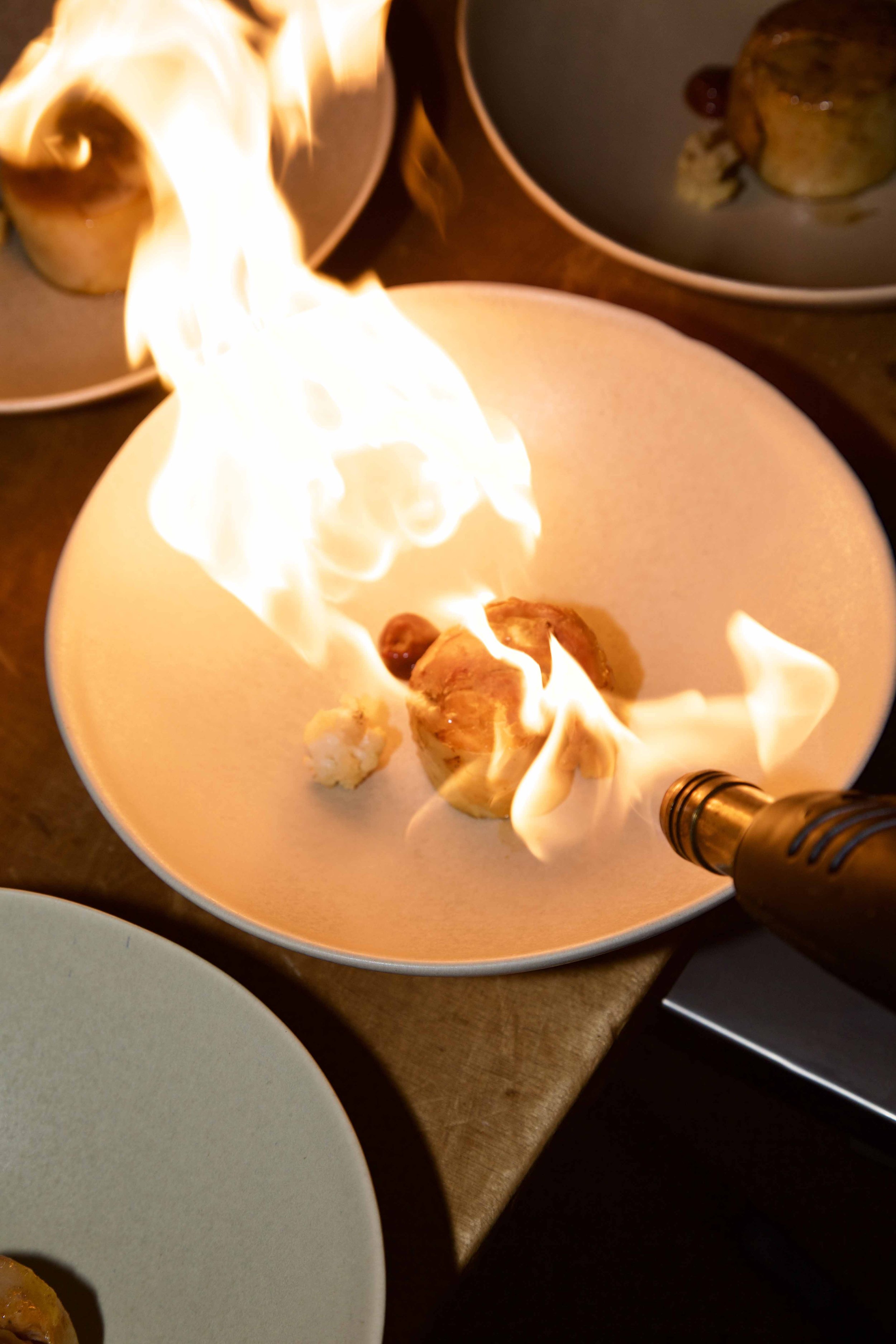 A plate with a dessert being flambéed with a flame from a torch, surrounded by other plates with similar desserts.