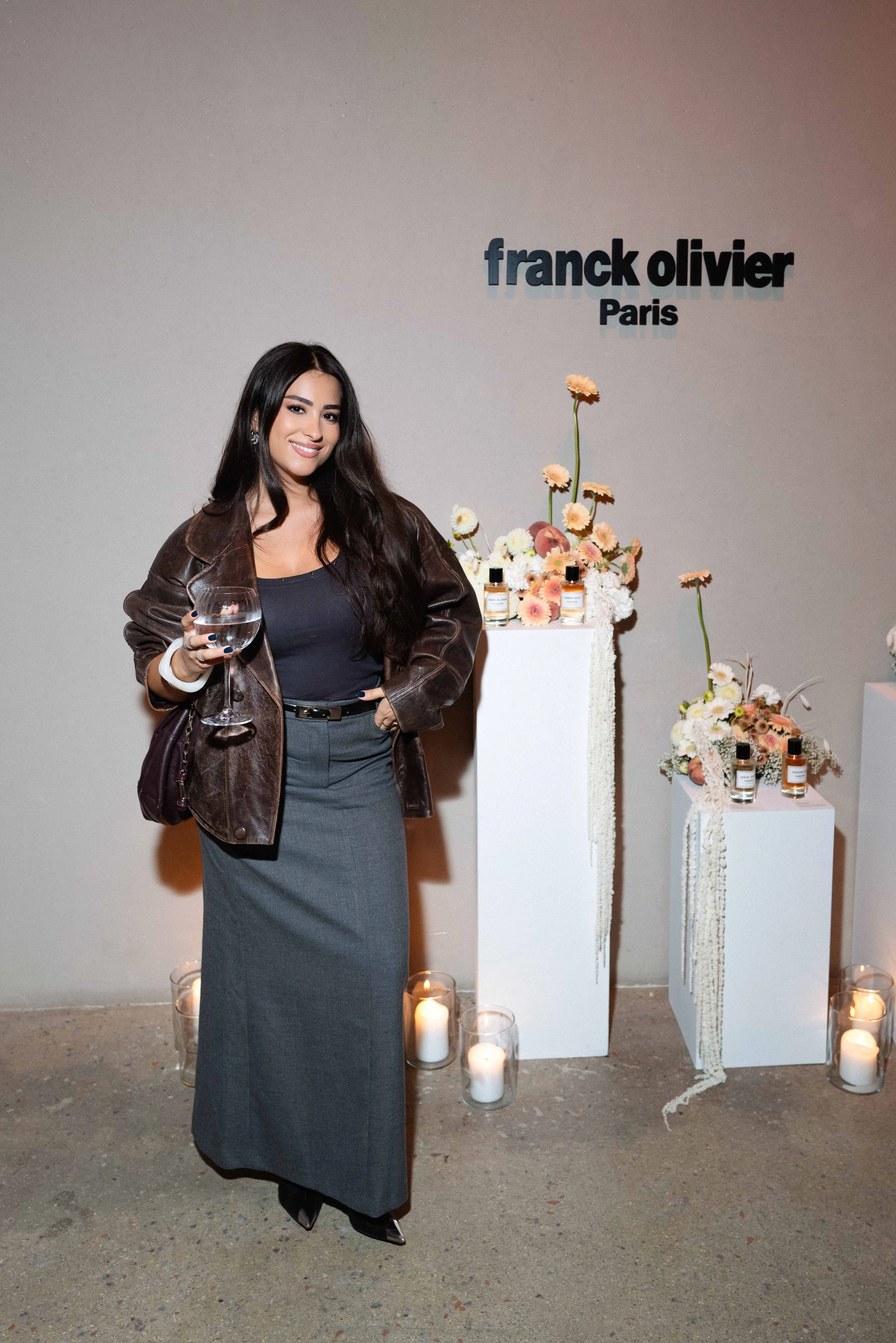 A woman with long dark hair, wearing a black top, gray skirt, and brown leather jacket, holding a glass of water, standing in front of a display with flowers and perfumes at an event for Franck Olivier in Paris.