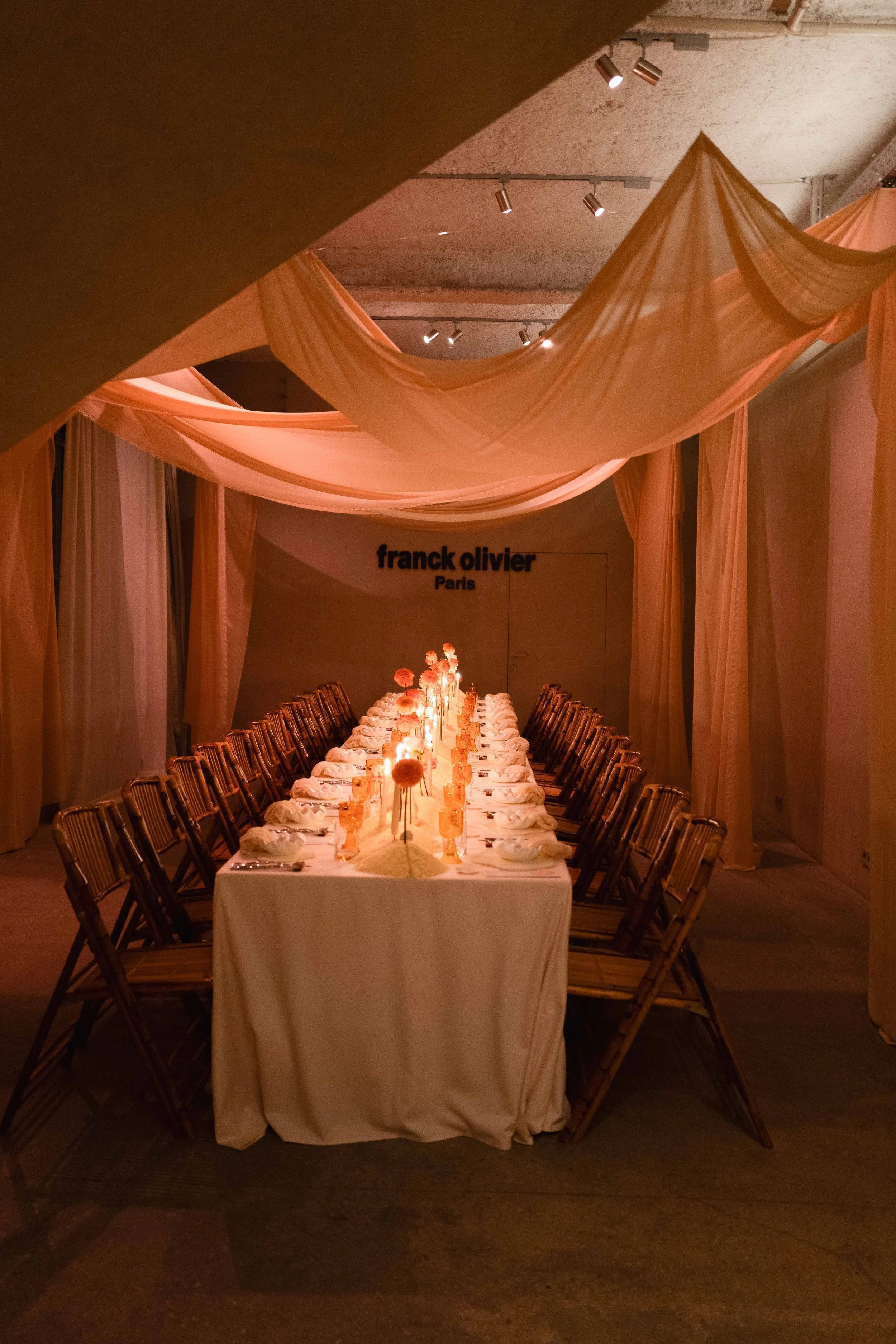 A long dinner table set for a fancy event with pink flowers and lit candles, surrounded by chairs, inside a dimly lit room with pink drapery and a "Franck Olivier Paris" sign on the wall.