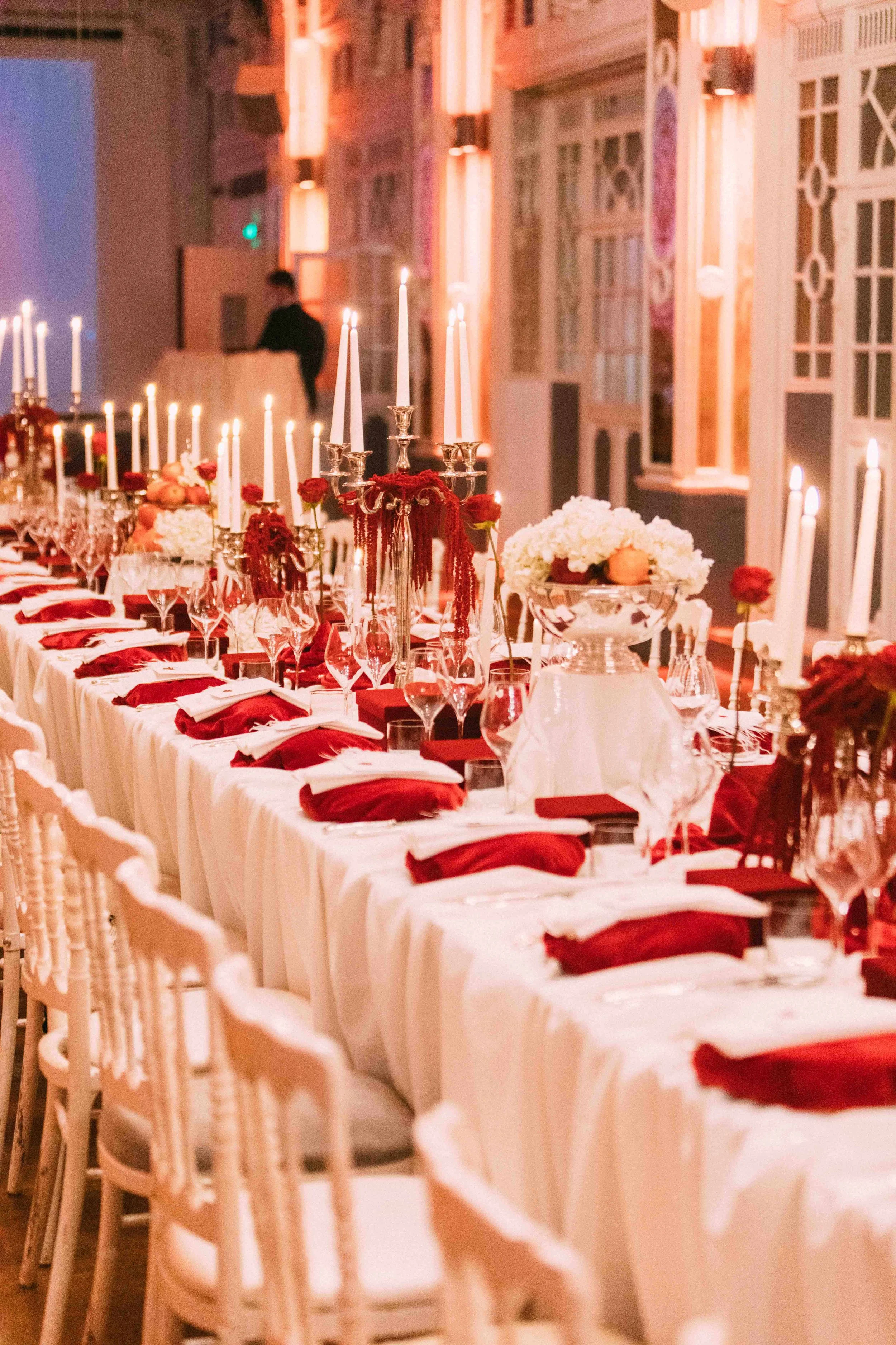 A long banquet table decorated with red and white floral centerpieces, red napkins, white tablecloth, and tall lit candles in a beautifully decorated room for an event or celebration.