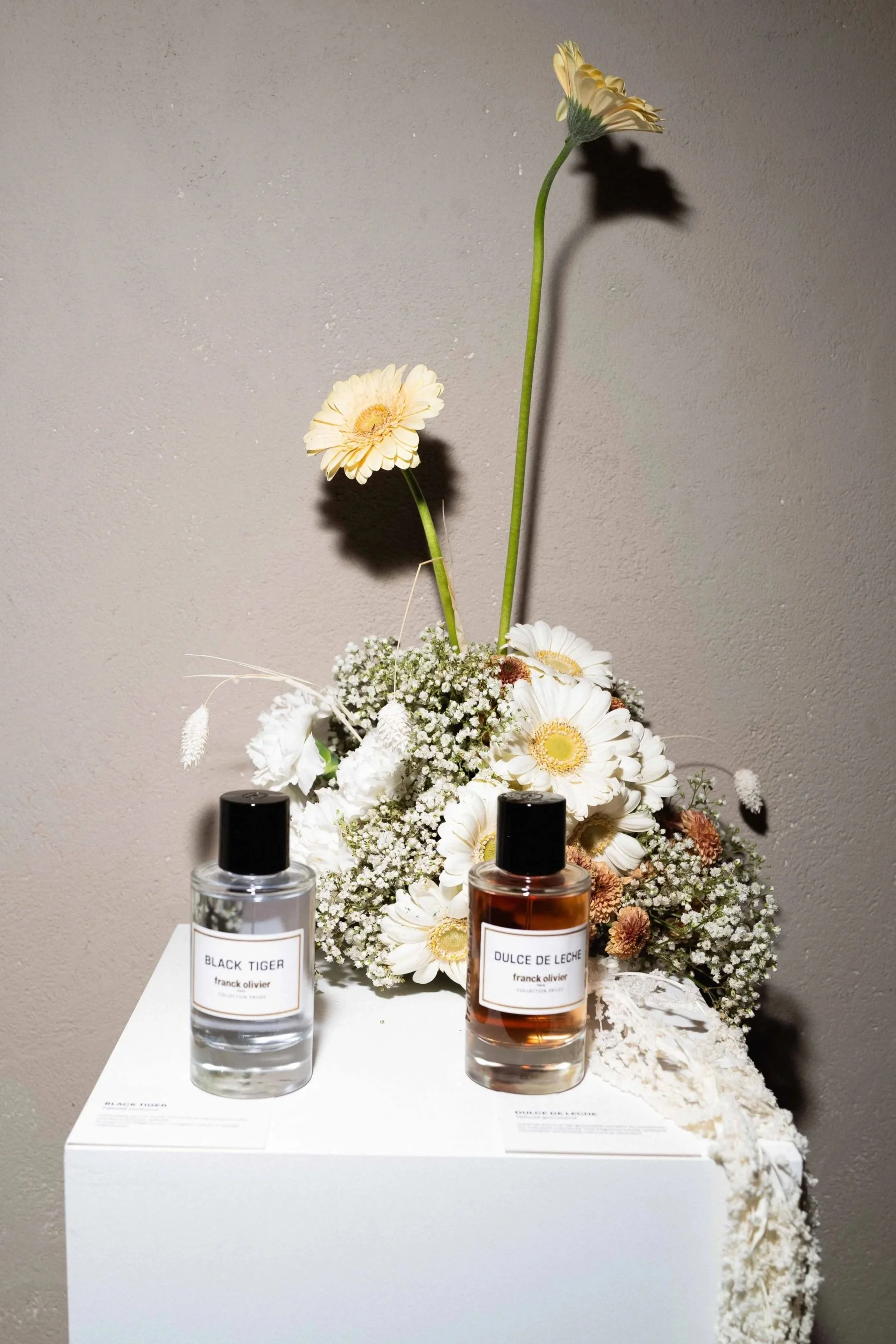 Display of two perfume bottles, one labeled 'Black Tiger' and the other 'Dulce de Leche,' in front of a white floral arrangement with daisies and baby's breath, set on a white pedestal against a gray wall.