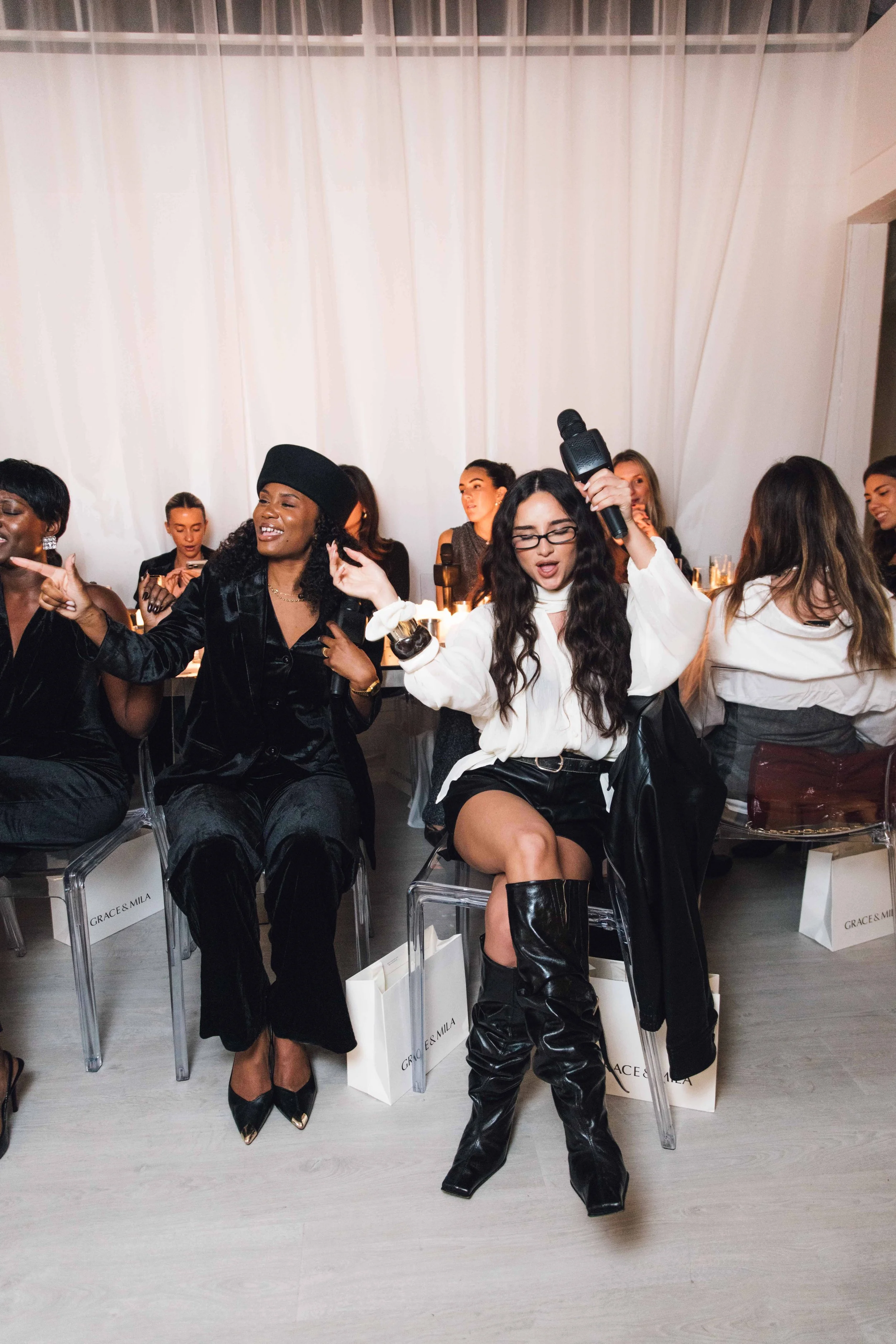 Women sitting in a row at a fashion event, some with microphones, surrounded by gift bags, with a white curtain backdrop.