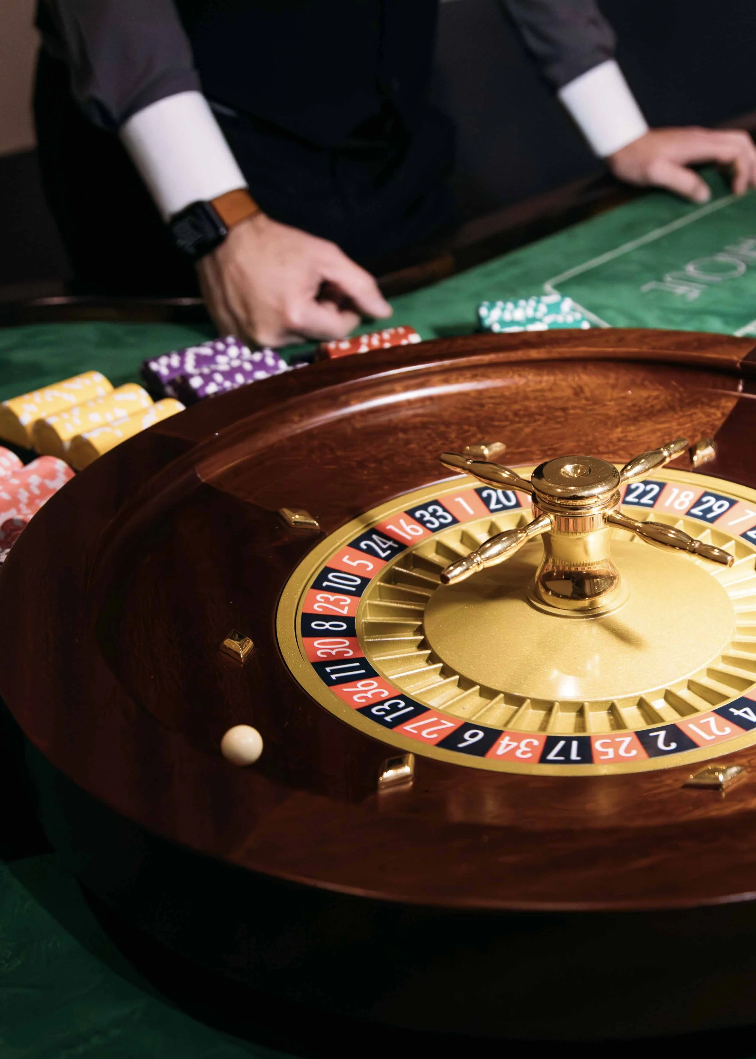 A roulette wheel with a white ball, casino chips, and a dealer in the background at a casino table.