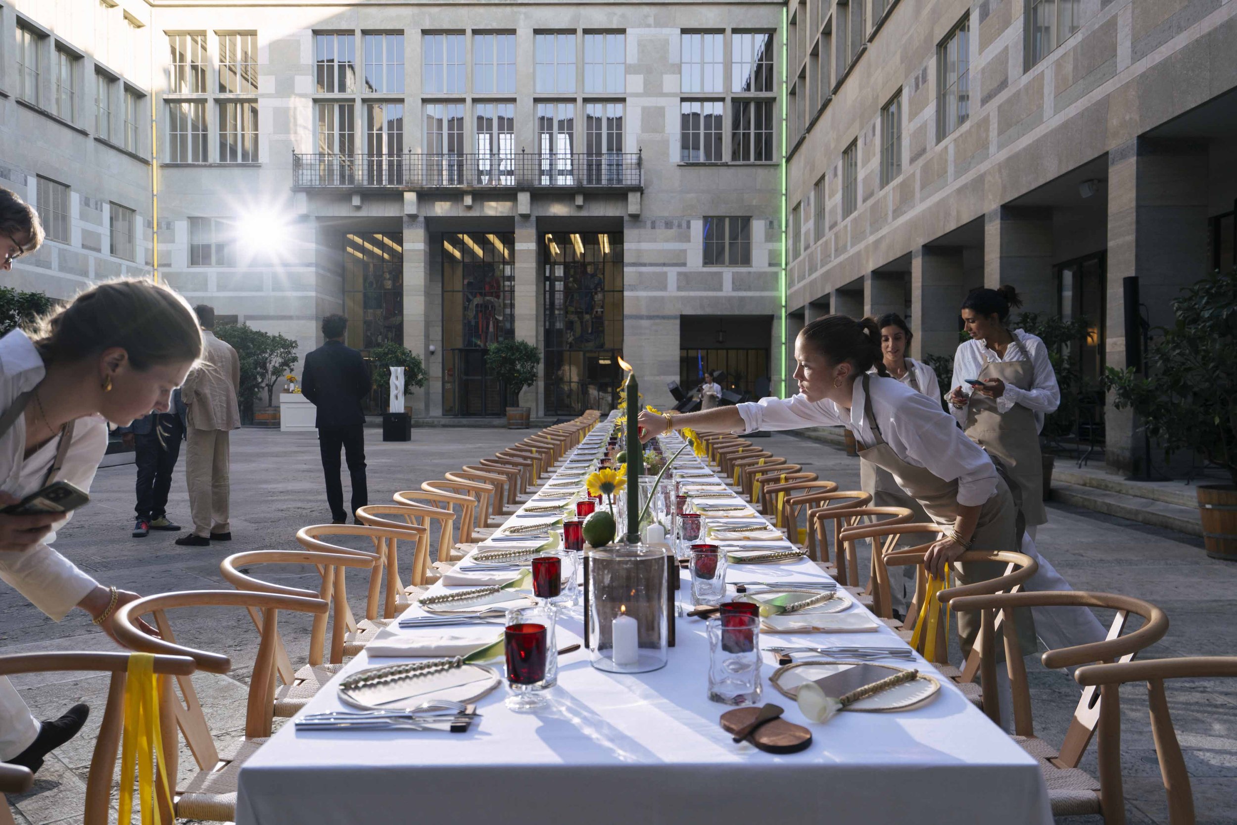 A long outdoor dining table set for a formal event in a courtyard of a large stone building, with servers preparing the table. The table is decorated with centerpieces, candles, and place settings.