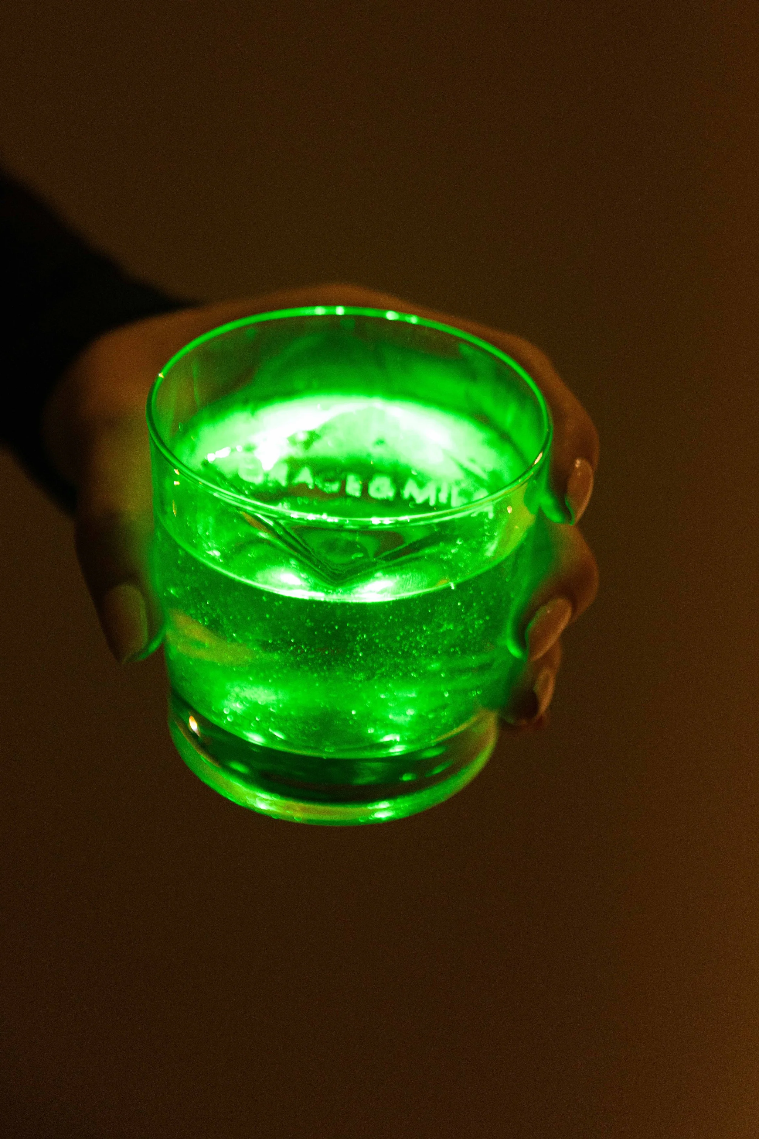 Person holding a glass of a glowing green drink in a dimly lit setting.